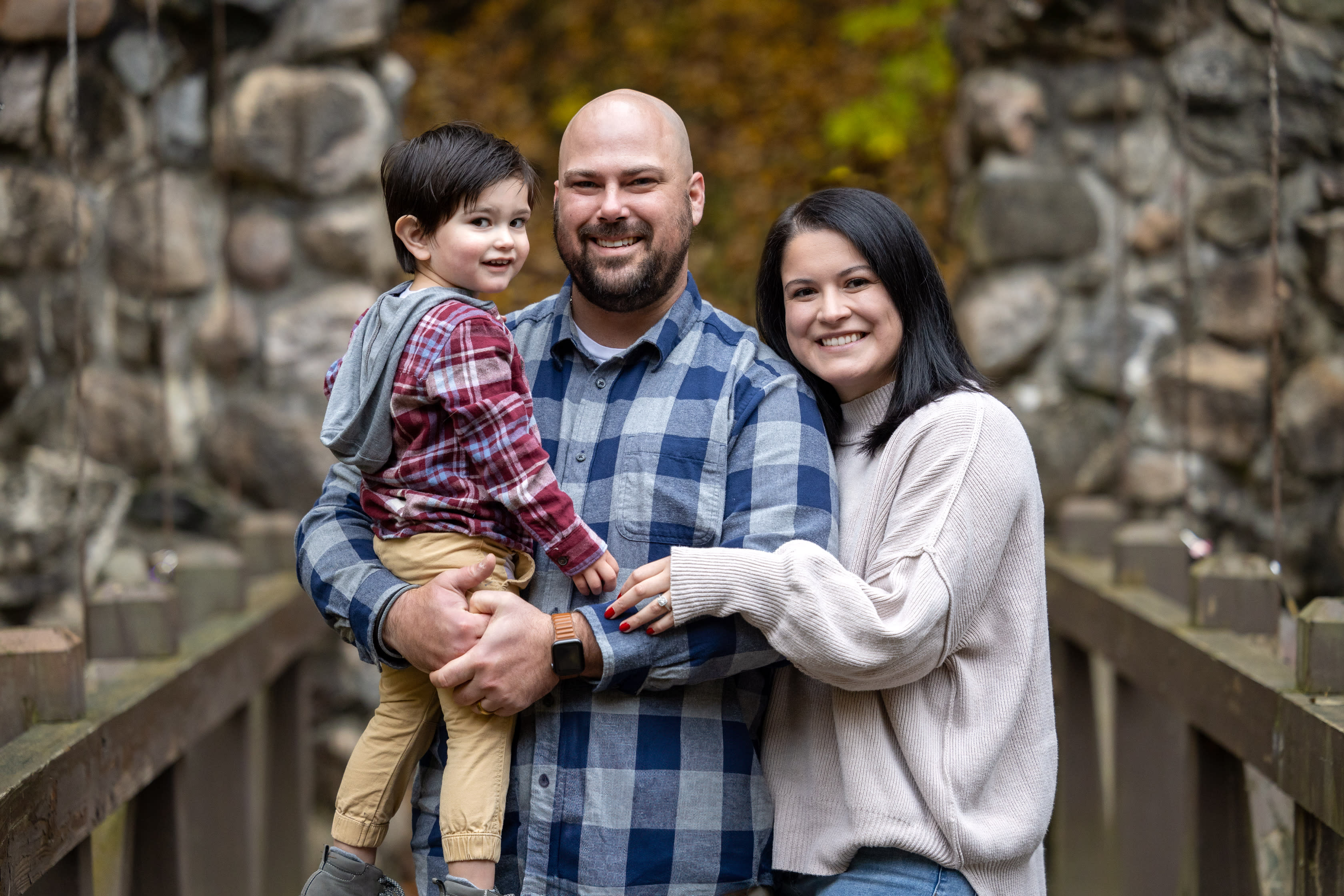 A smiling family of three stands on a wooden bridge with stone walls in the background. The man in the middle, showcasing remarkable work-life balance, holds a young boy in one arm. The woman stands to the right, hugging the man. The boy wears a red shirt; the adults wear blue plaid and a light sweater, respectively.