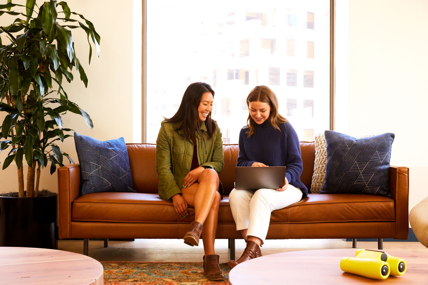Two people sit together on a brown leather couch in a modern office space with a large window and potted plant. One person is wearing a green jacket and shorts, while the other wears a blue sweater and white pants. They are both smiling.