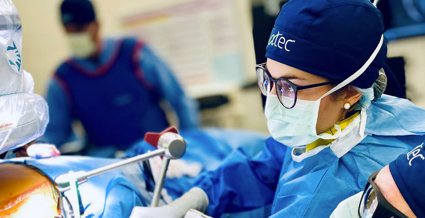 Two medical professionals in a surgical setting. The foreground shows a person in glasses and a surgical mask, focusing carefully. Both wear blue scrubs, caps, and protective gear. 