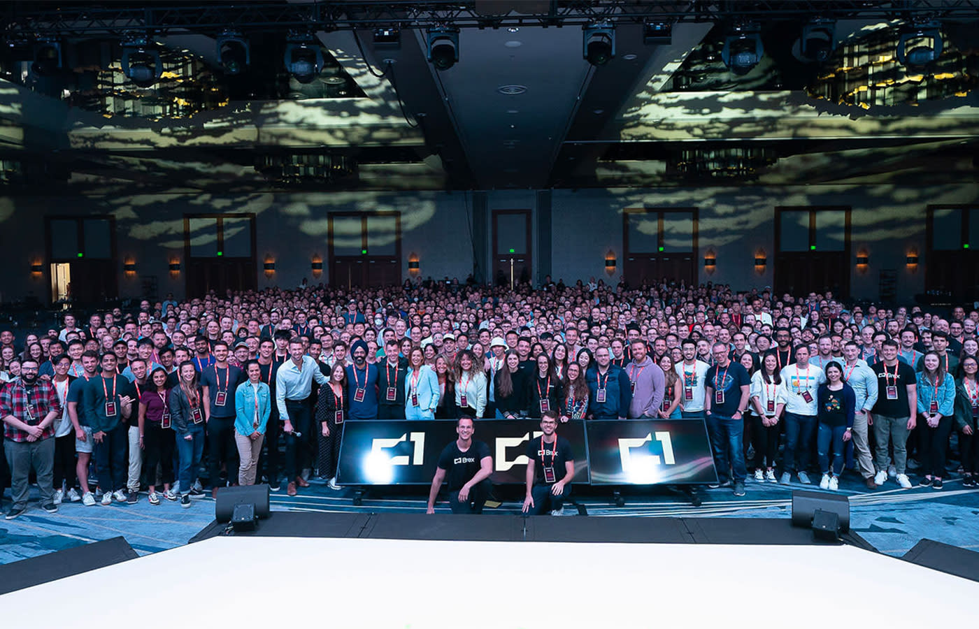A large group of people are gathered in a spacious, indoor venue for a group photo. Many individuals wear name tags, and two people are seated in front of a stage setup with the letter "F1" displayed. The background features decorative wall projections and lighting.