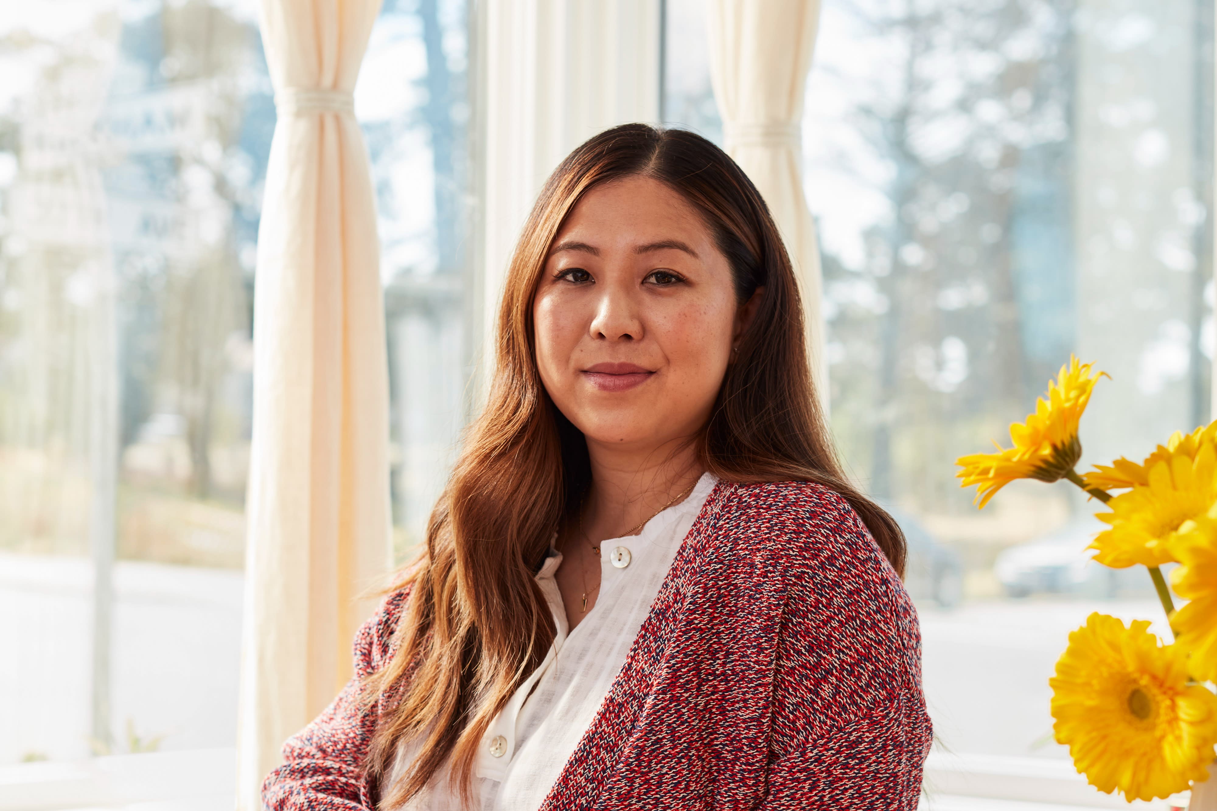 A woman with long, wavy brown hair wearing a white blouse and red cardigan sits by a window with sheer curtains. She gazes softly at the camera. Bright yellow flowers in a vase are visible on her right side. The serene setting puts one at ease, evoking thoughts of employee leave compliance under the CFRA.