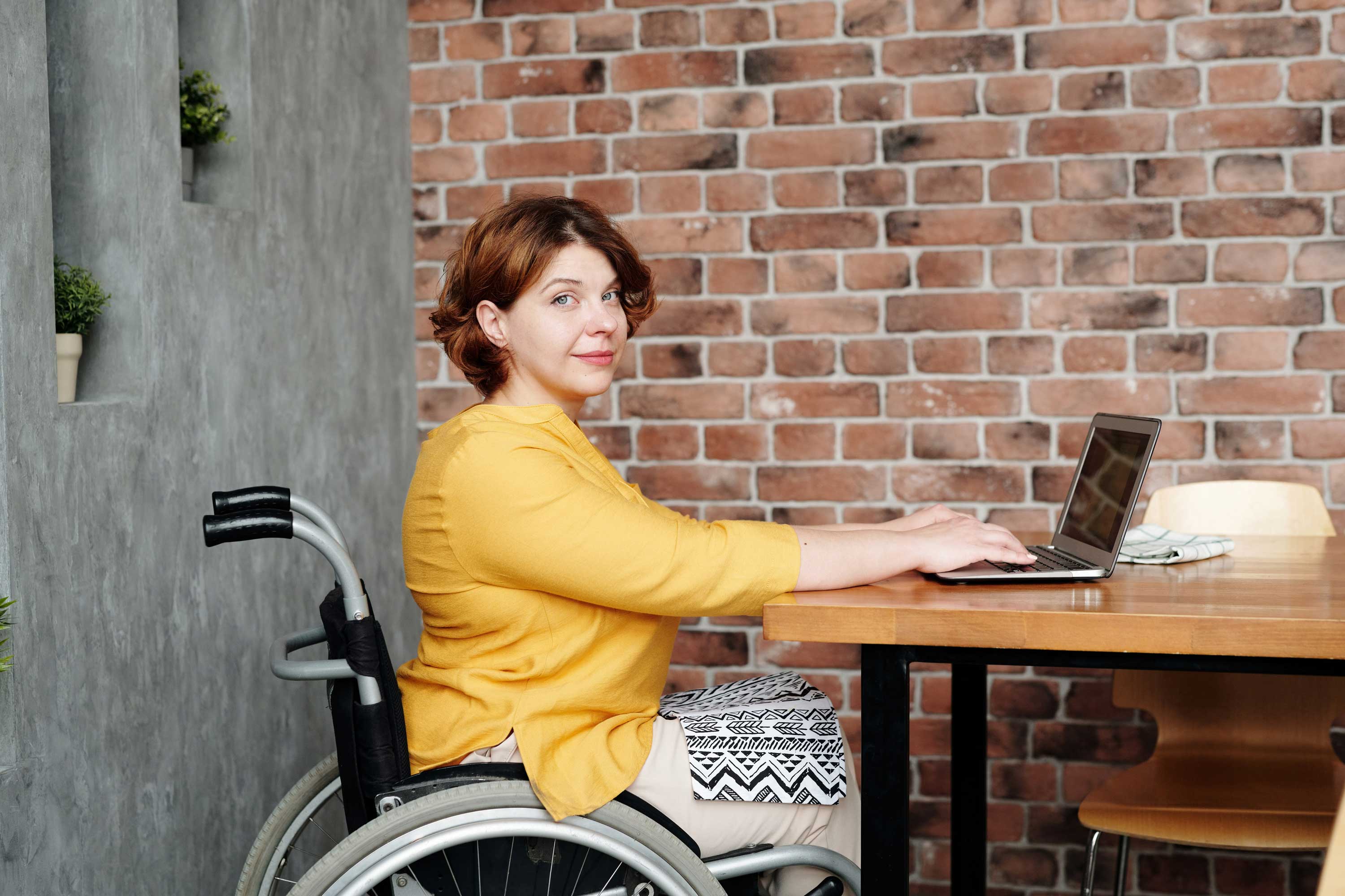 A woman with short brown hair sits in a wheelchair, wearing a yellow top and patterned skirt. She works on a laptop at a wooden table, backed by a brick wall and gray decor with small plants on the shelves. She looks at the camera and smiles slightly, advocating for disability benefits awareness.