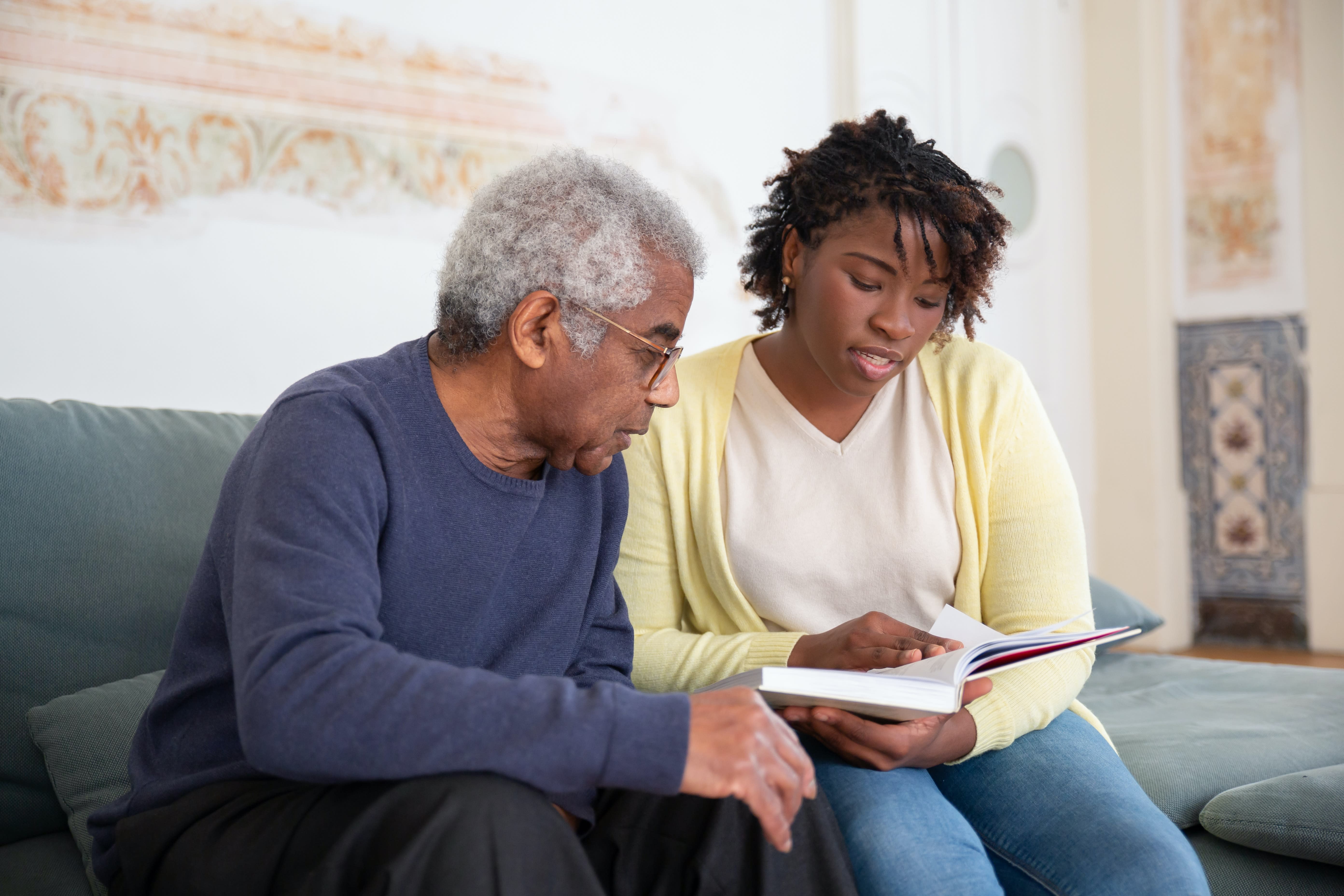 A young woman and an older man sit on a couch together. The young woman, on the right, is holding a book and pointing to a page while they both read. The older man, on the left, attentively looks at the book. They are in a cozy, well-lit room discussing Rhode Island TCI benefits.