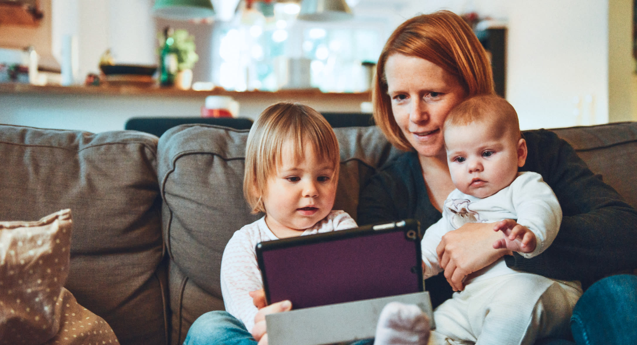 A woman with red hair is sitting on a couch holding a baby and a young child. The young child is holding a tablet, and they all appear to be looking at the screen. The background shows a bright living room with various household items, providing a snapshot of quality family time during FMLA job-protected leave.