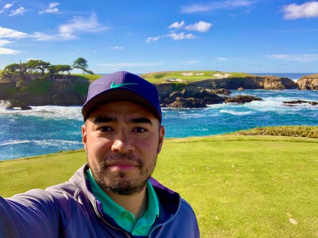 Chris is in a purple cap and gray jacket with a teal collar as he takes a selfie on a lush, green golf course with the ocean and rocky cliffs in the background under a bright blue sky.