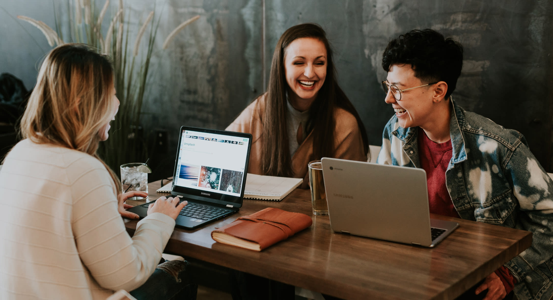 Three smiling individuals are seated around a wooden table, each with an open laptop, discussing retention strategies. Two women and one person with short hair and eyeglasses are engaged in conversation. A plant and a notebook are also on the table. The background features a textured wall.