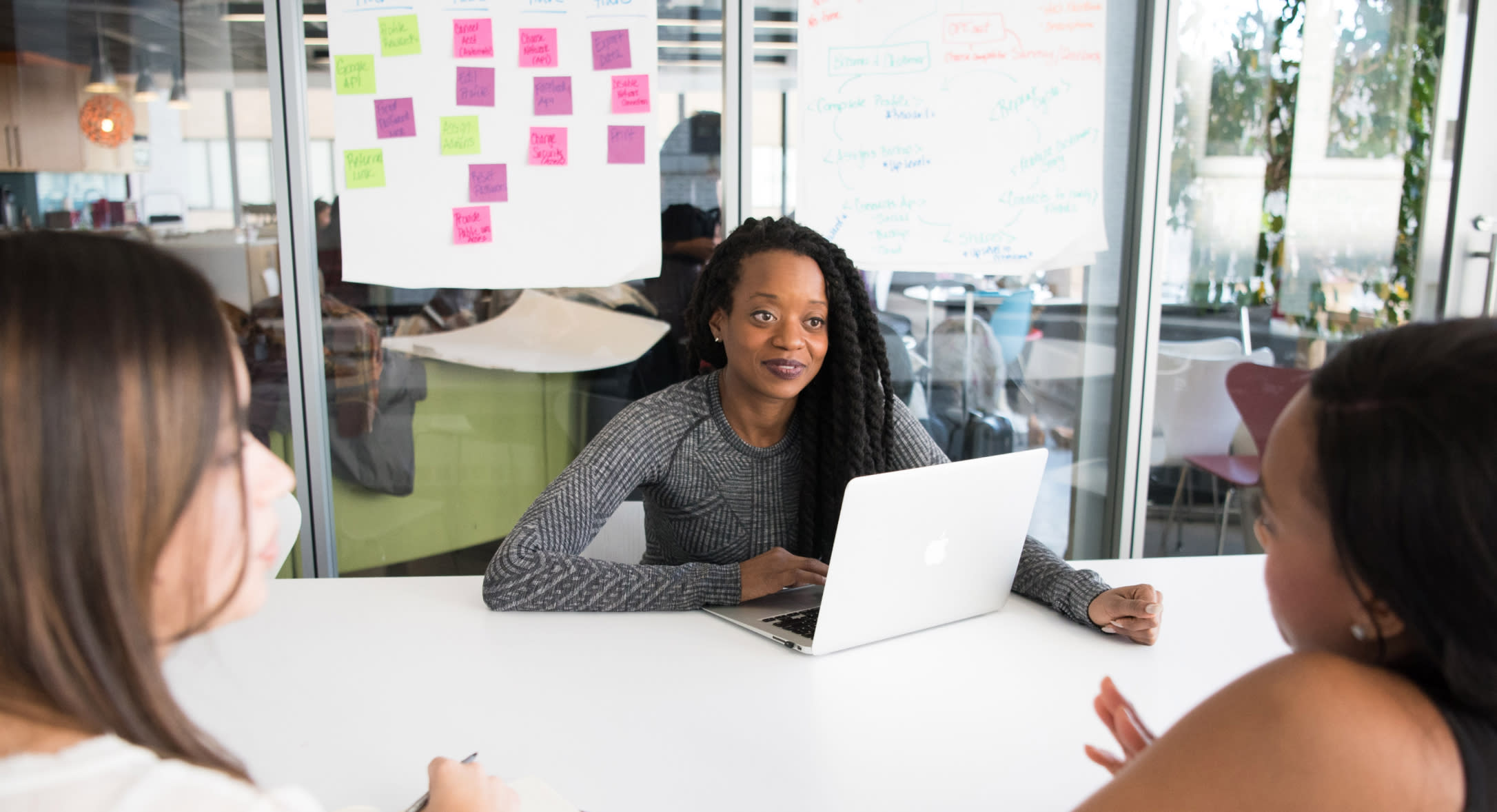 A woman with long braids sits at a table with a laptop, engaging in a discussion with two colleagues about leave compliance. Colorful sticky notes and a whiteboard with writing can be seen in the background, indicating a collaborative and creative workspace.