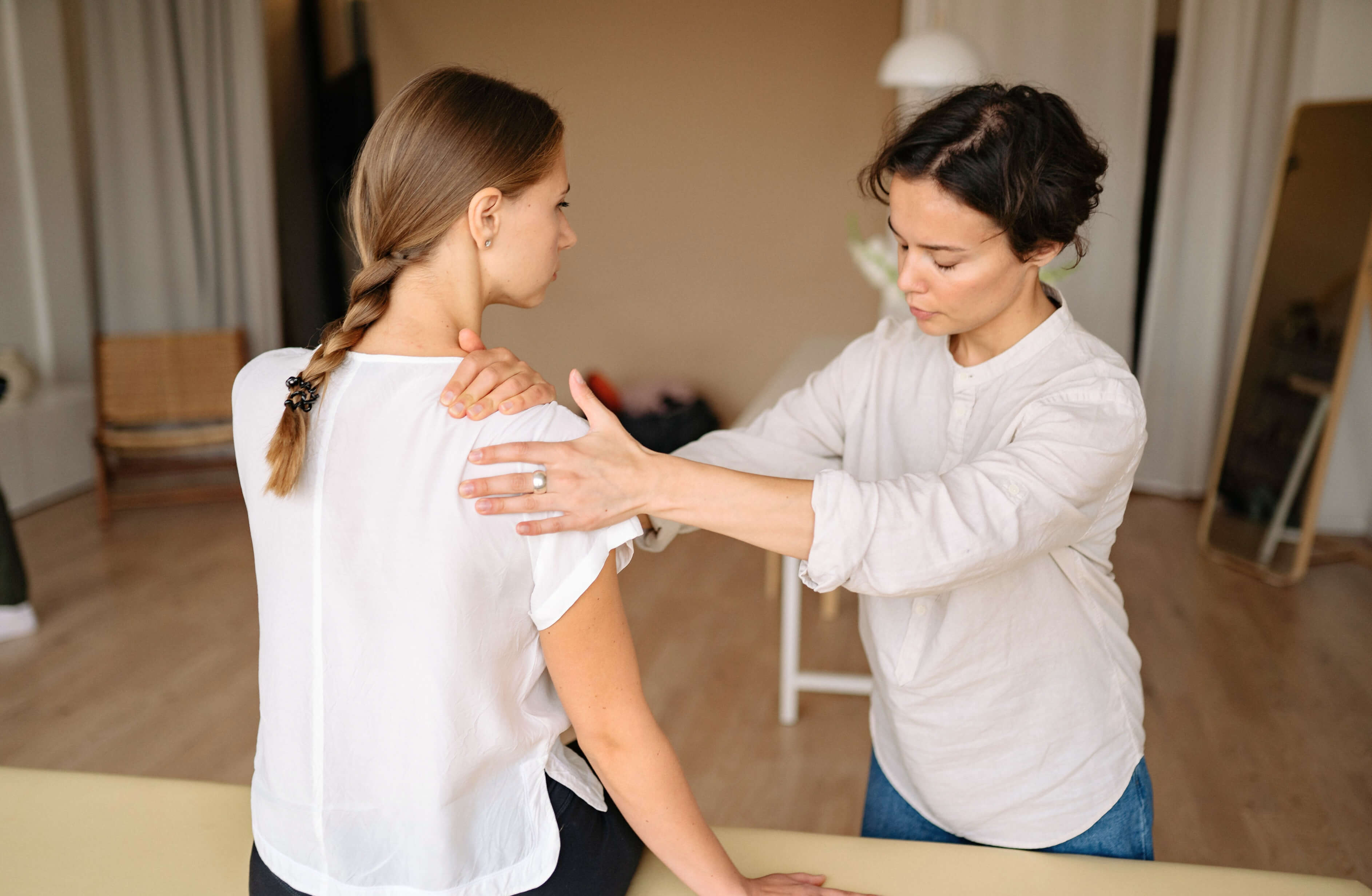 A woman with a braid sits while another woman with short hair performs a physical therapy or chiropractic adjustment on her shoulder in a well-lit room, potentially discussing disability insurance compliance. The person performing the therapy uses both hands to apply gentle pressure.