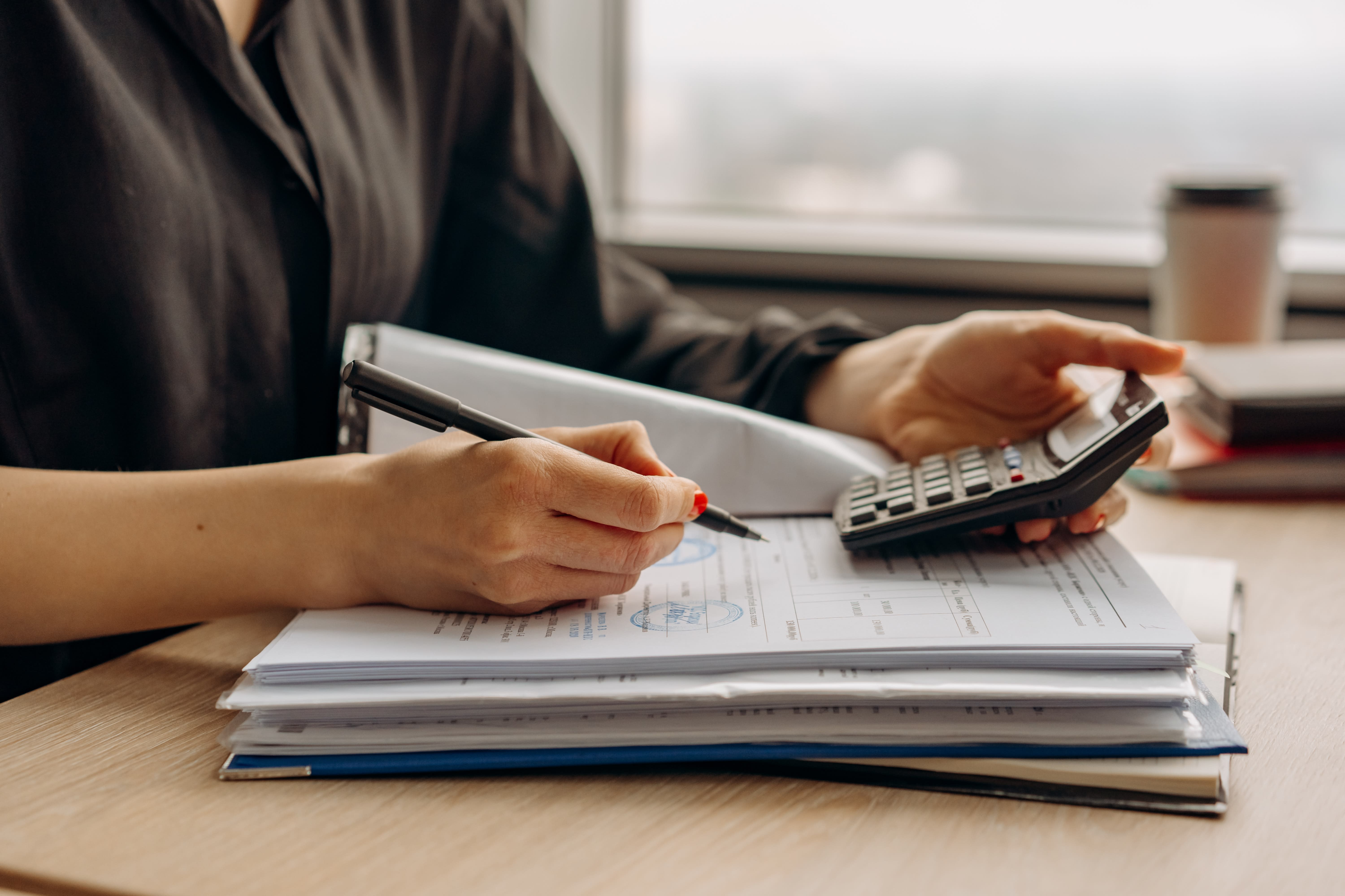 A person in black attire is seated at a desk, using a calculator with one hand while writing on a document about D.C. disability insurance with the other. The desk has stacks of paper, a binder, and a coffee cup in the background, suggesting an office or workspace