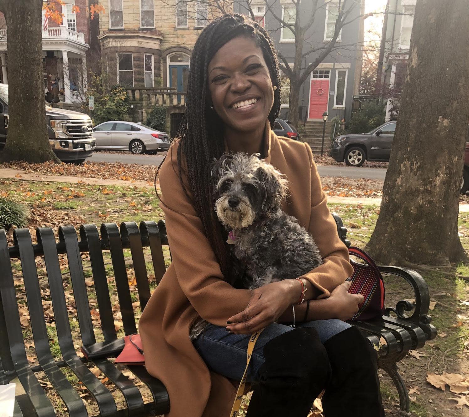 A woman with long braided hair is sitting on a park bench, smiling and holding a small, fluffy dog. Wearing a tan coat and blue jeans, she looks composed. The background shows trees, parked cars, residential buildings, and fallen leaves.