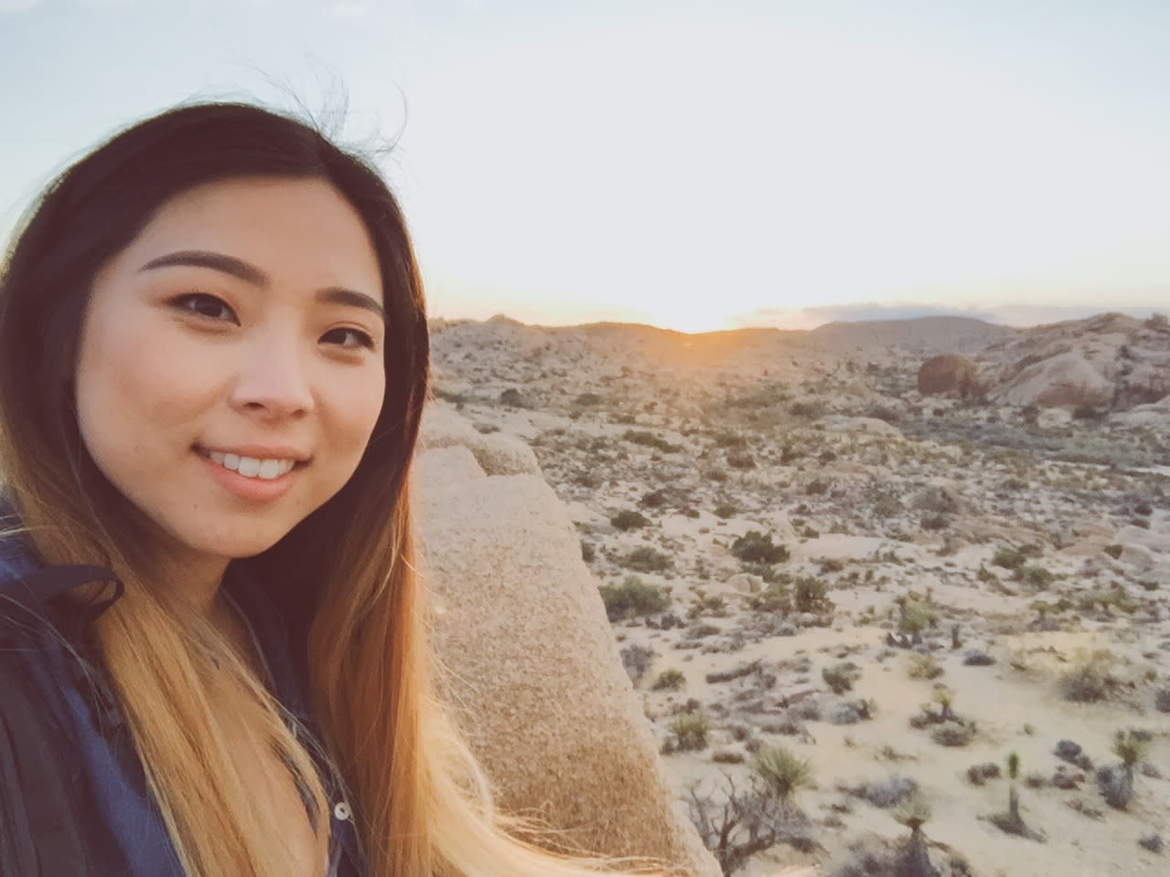 Erika, with long hair flowing, takes a selfie outdoors in the desert landscape at sunset. She's smiling, with rocky terrain and sparse vegetation visible in the background. The clear sky and soft, warm light illuminate the scene beautifully.