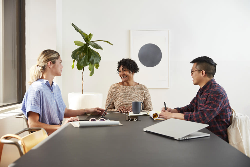 Three people sit around a conference table engaging in a discussion. One person on the left wears a light-colored shirt, another in the center wears a beige top, and the person on the right wears a checkered shirt. Amidst their conversation about Exabeam, a laptop, books, and a potted plant are visible.