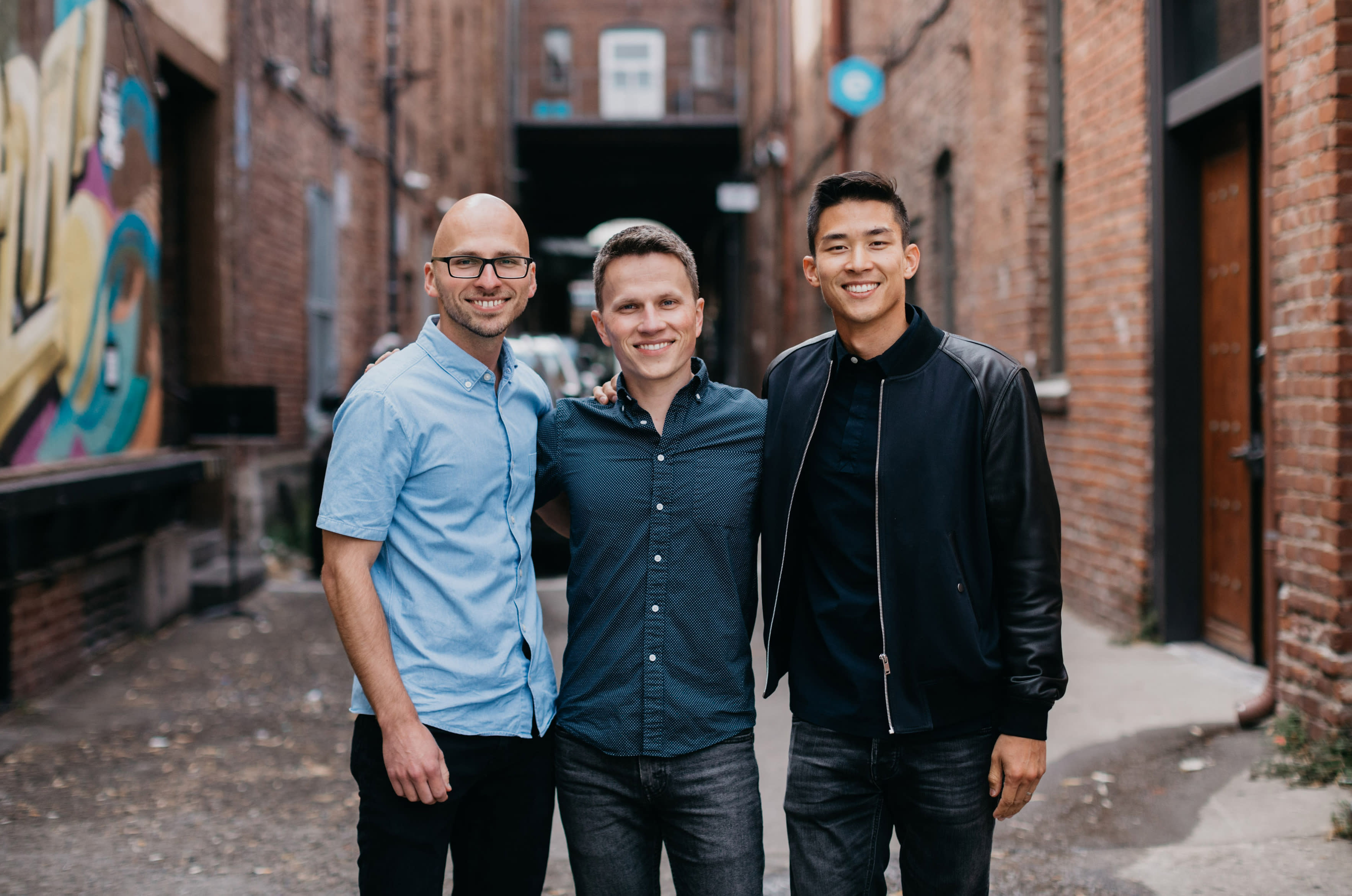 Three men are standing close together and smiling in a narrow, brick alleyway. With arms around each other, one man in a light blue shirt, another in dark blue, and the last wearing a black jacket over a dark shirt exude camaraderie that reminds one of seamless teamwork and HR efficiency.