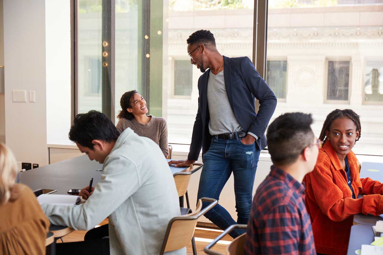 A group of diverse people is gathered in a modern office space with large windows, reflecting the exceptional employee experience at one of the Best Small Workplaces. Four are seated at a rectangular table, engaged in conversation and note-taking. One person, standing and leaning on the table, is talking with another.