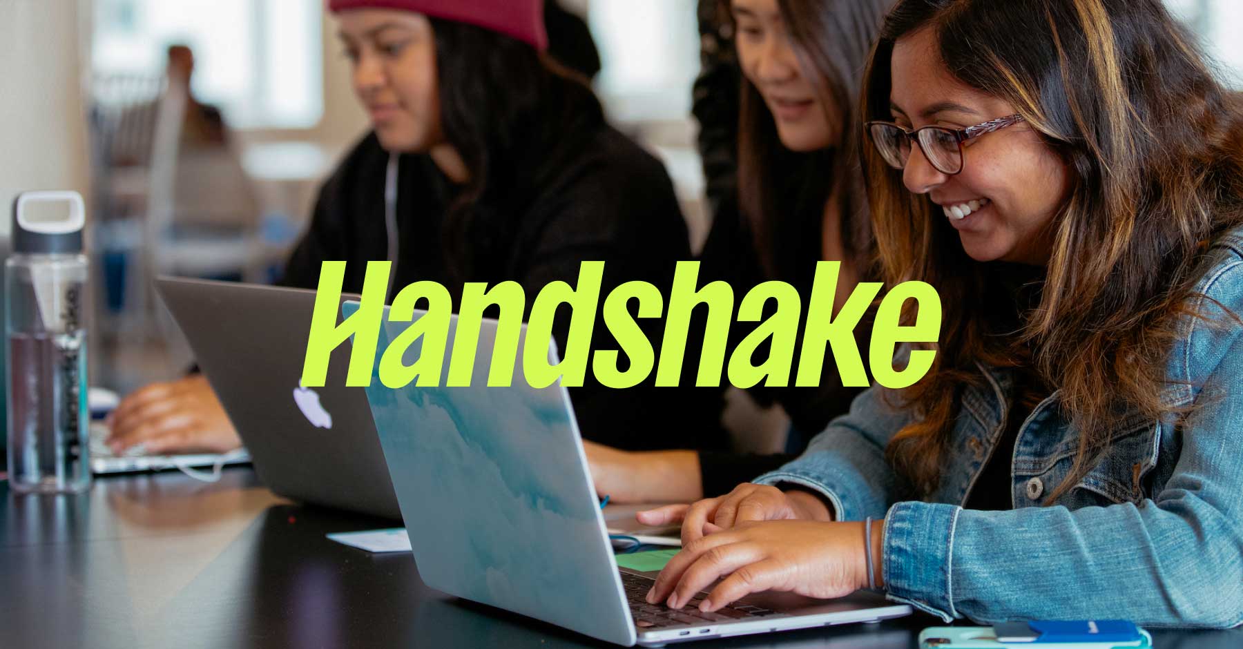 Three people are sitting at a table working on laptops, appearing focused and engaged. The word "Handshake" is prominently displayed in bold yellow-green text across the image. A water bottle is visible on the table to the left.