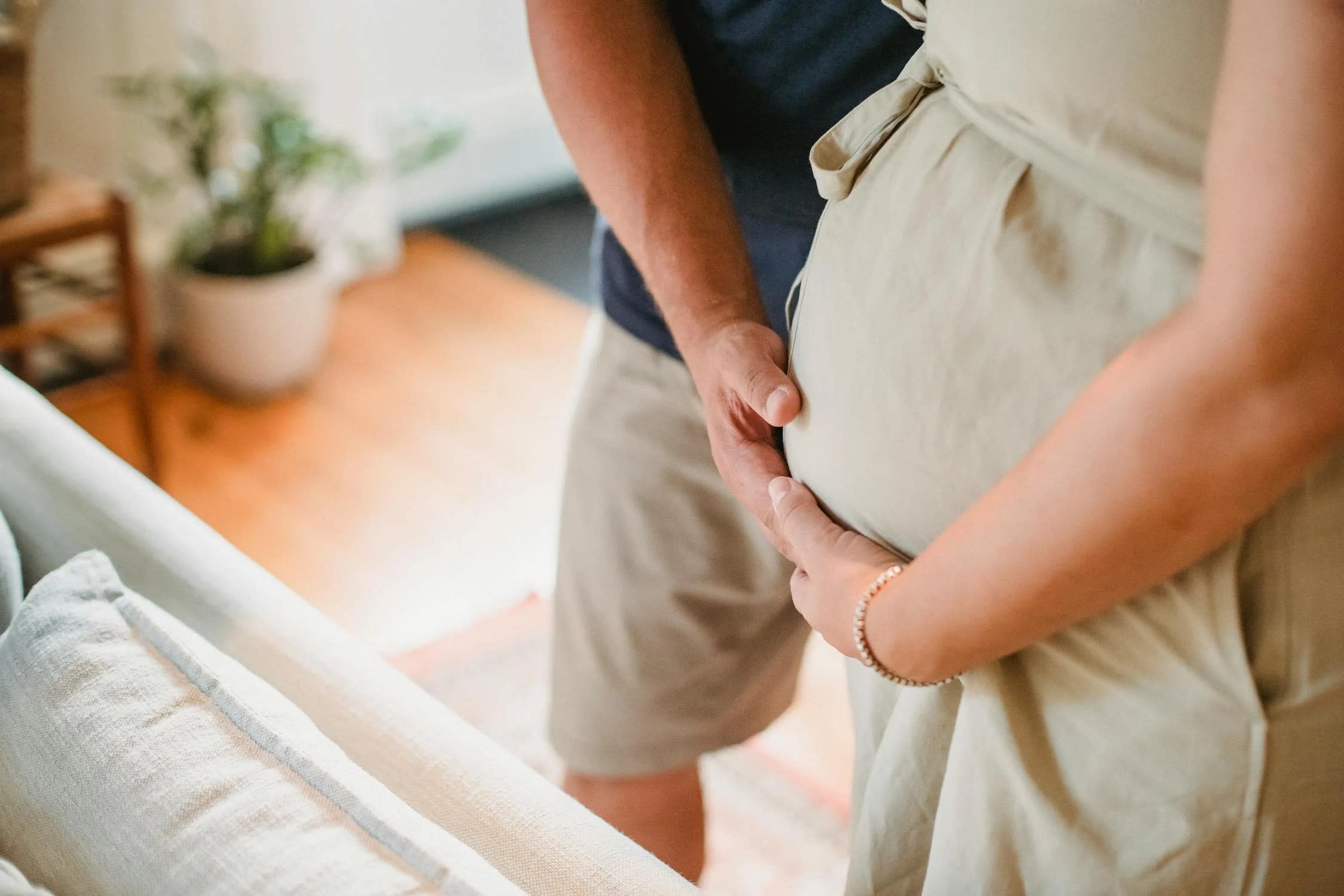A close-up of a couple indoors, with the man's hands gently placed on the pregnant woman's belly. Both are dressed in casual summer clothing. The background features a blurred-out potted plant and part of a wooden chair, symbolizing the need for reasonable accommodations for expectant parents in the workplace.