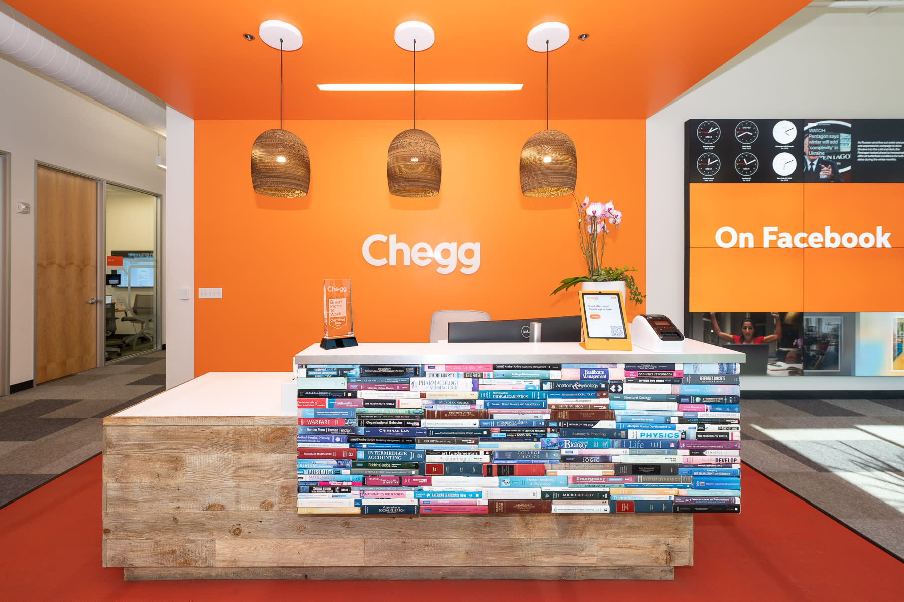 A reception area with an orange and white color scheme features a desk adorned with stacked books. Above, three woven pendant lights hang, while a sign reads "Chegg." To the right, a digital screen highlights "Parental Leave" benefits on Facebook.
