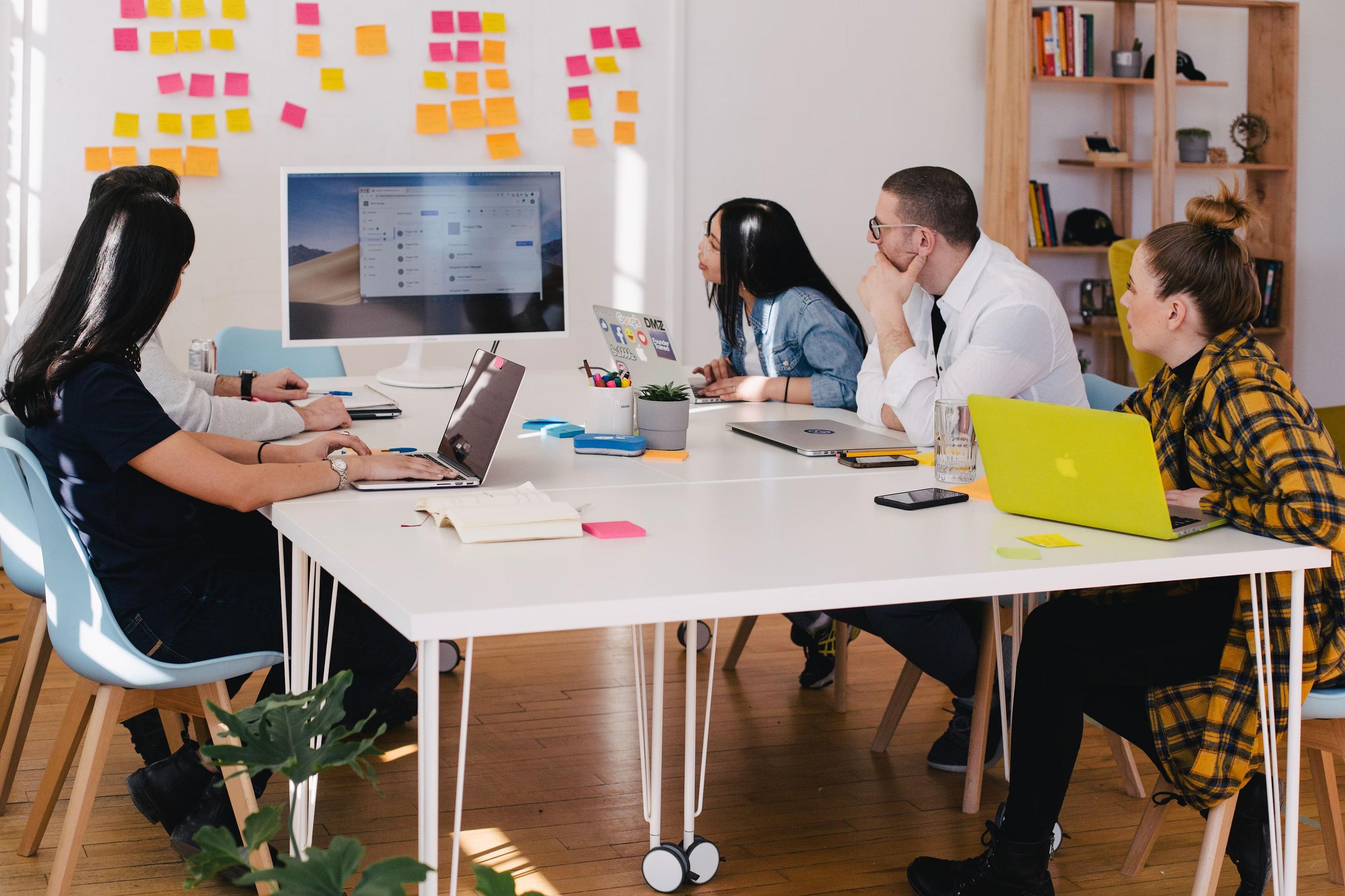 A group of five people, possibly part of the Sparrow team, sits around a table in an office setting, intently focused on a large computer monitor displaying a document. Laptops and colorful sticky notes are scattered across the table. Shelves and more sticky notes adorn the background.