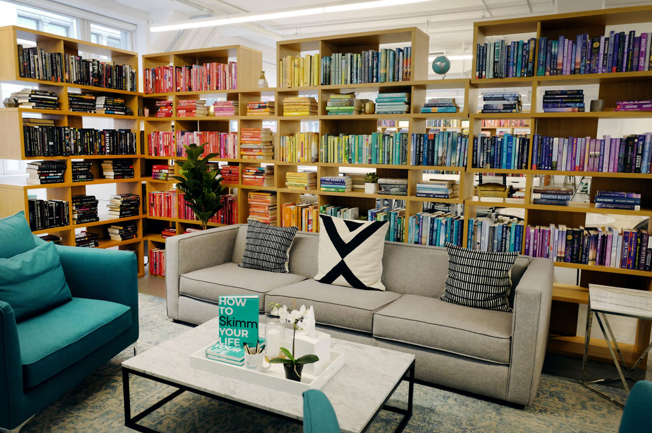 A cozy reading area features a gray couch and two teal chairs around a marble coffee table. The bookshelves display books organized by color, adding a rainbow effect. A green book titled "How to Skimm Your Life" lies on the table, alongside pamphlets on Sparrow leave solutions.