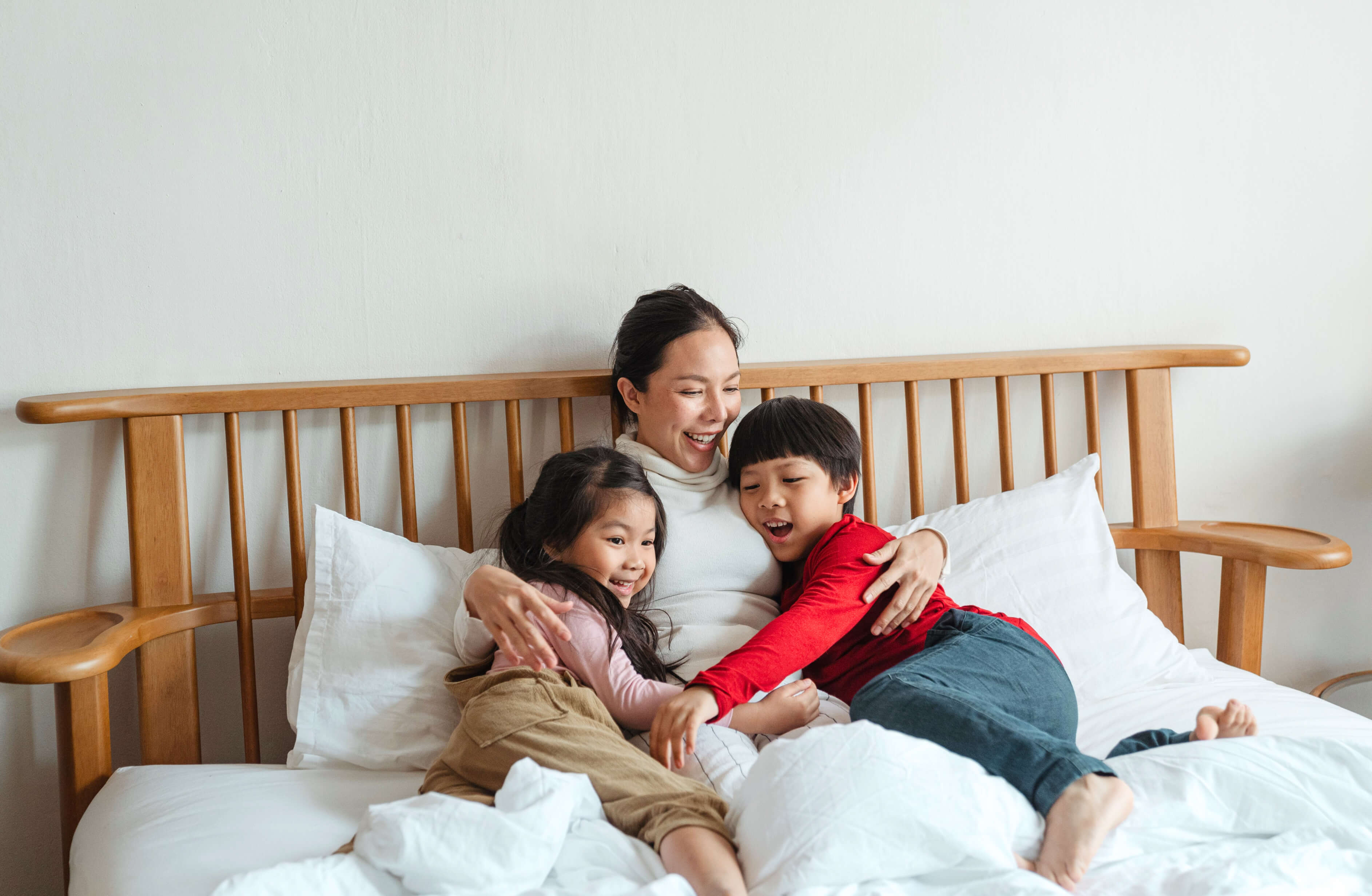 A woman sits on a bed, smiling and hugging two children on either side of her. The child on the left is wearing a pink shirt and beige pants, while the child on the right is wearing a red shirt and dark pants. Thanks to paid time off in Illinois, they all appear to be laughing and enjoying their time together.