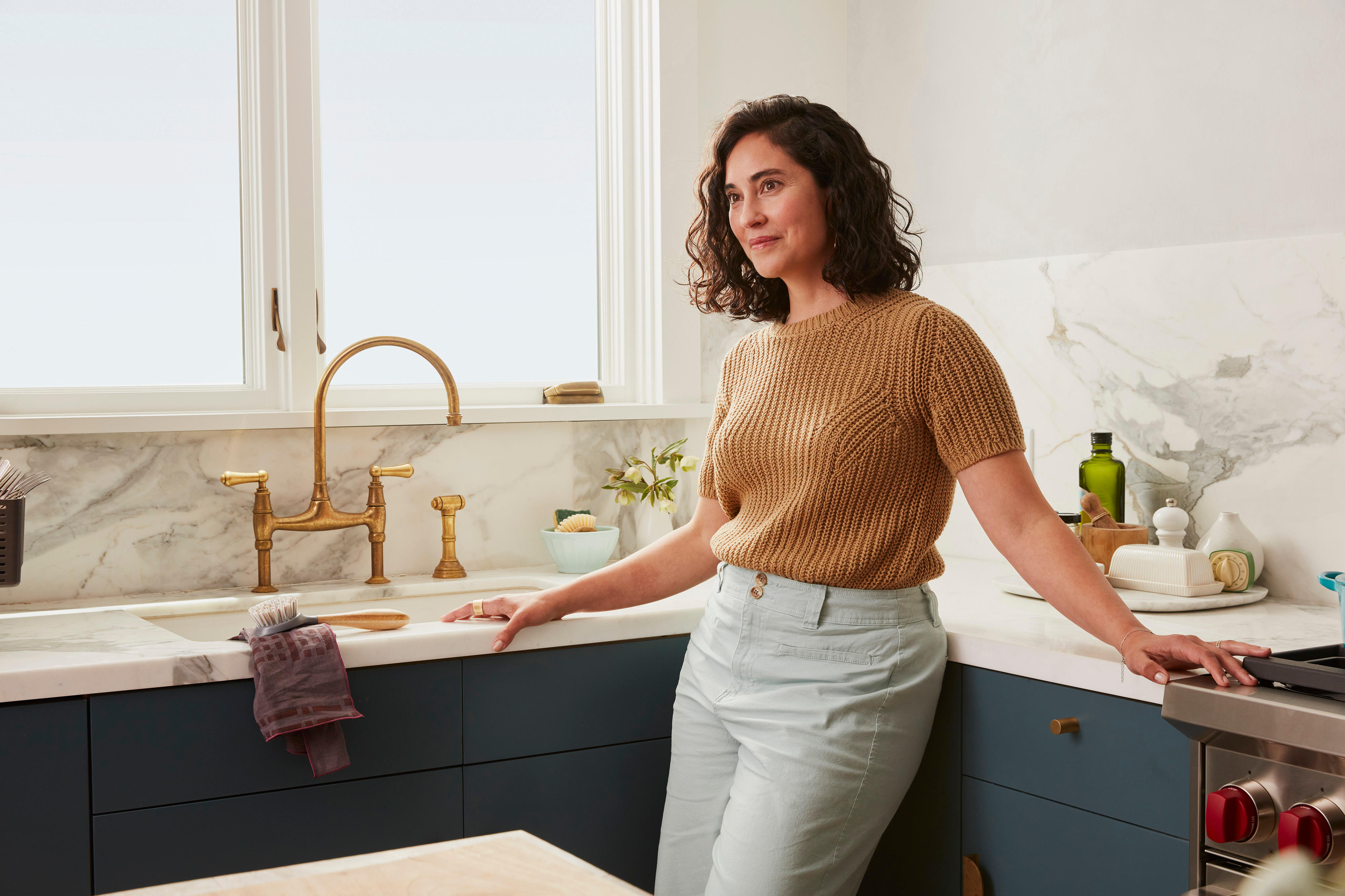 A woman with curly dark hair, wearing a tan knit sweater and light pants, leans against a marble countertop in a bright, modern kitchen. The kitchen features navy blue cabinets, a brass sink, and a window allowing natural light to fill the space while she thinks about Illinois S.B. 3120 concerning bereavement leave requirements.