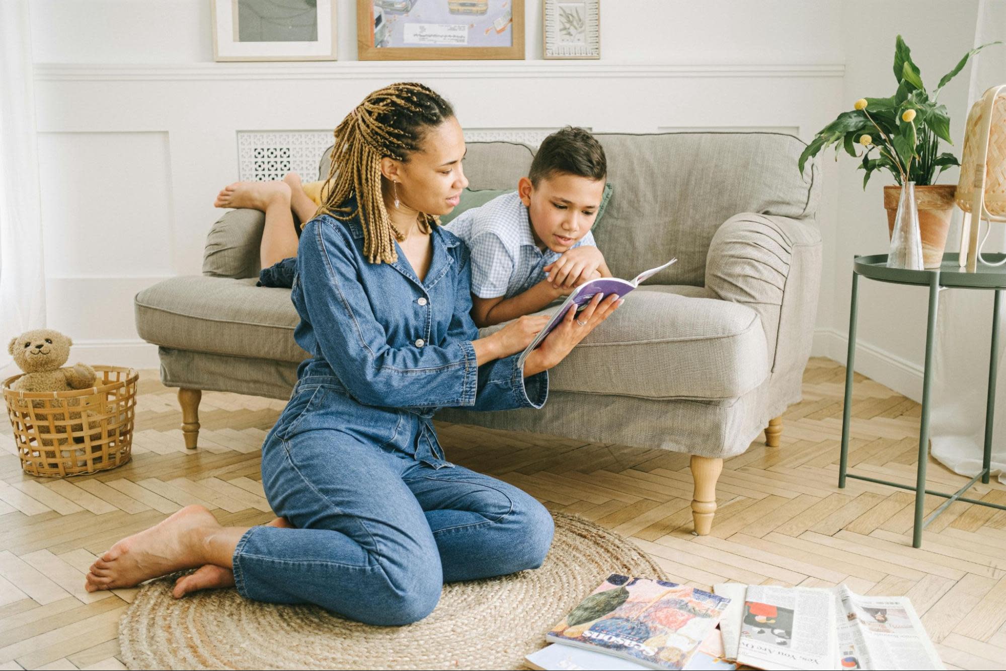 A woman with braided hair, wearing a denim jumpsuit, is sitting on a round woven rug, reading a book to a young boy. The boy is lying on a grey sofa, leaning on his elbows, attentively looking at the book. The setting exudes cozy vibes in their bright living room, reflecting the peace supported by Oregon Family Leave Act.