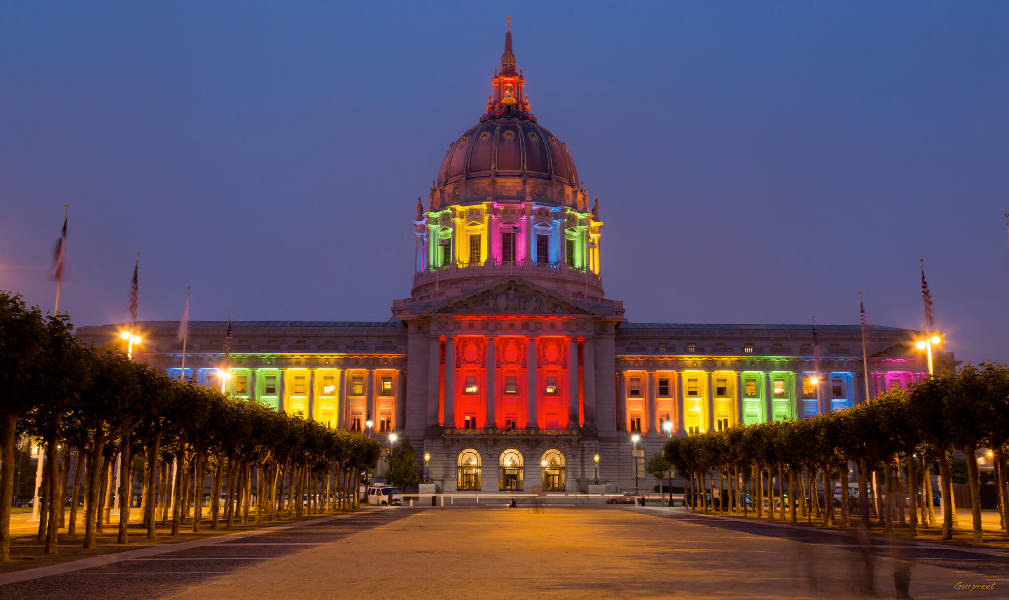 A grand building with a large dome is illuminated with vibrant rainbow colors at night, symbolizing LGBTQ+ pride. The structure is framed by lit trees along a walkway leading to the entrance, creating a festive and welcoming atmosphere.