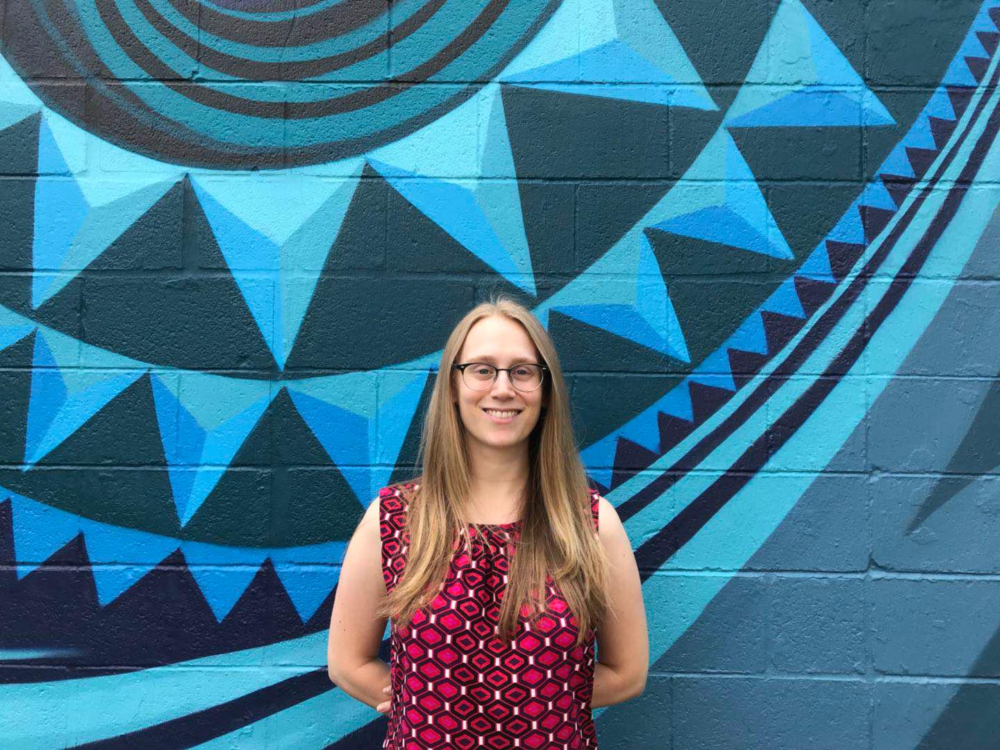 Emily, a member of the Sparrow Sales Team, is smiling and standing in front of a brightly colored geometric mural with shades of blue and black. With long hair and glasses, she wears a sleeveless red and black patterned top.