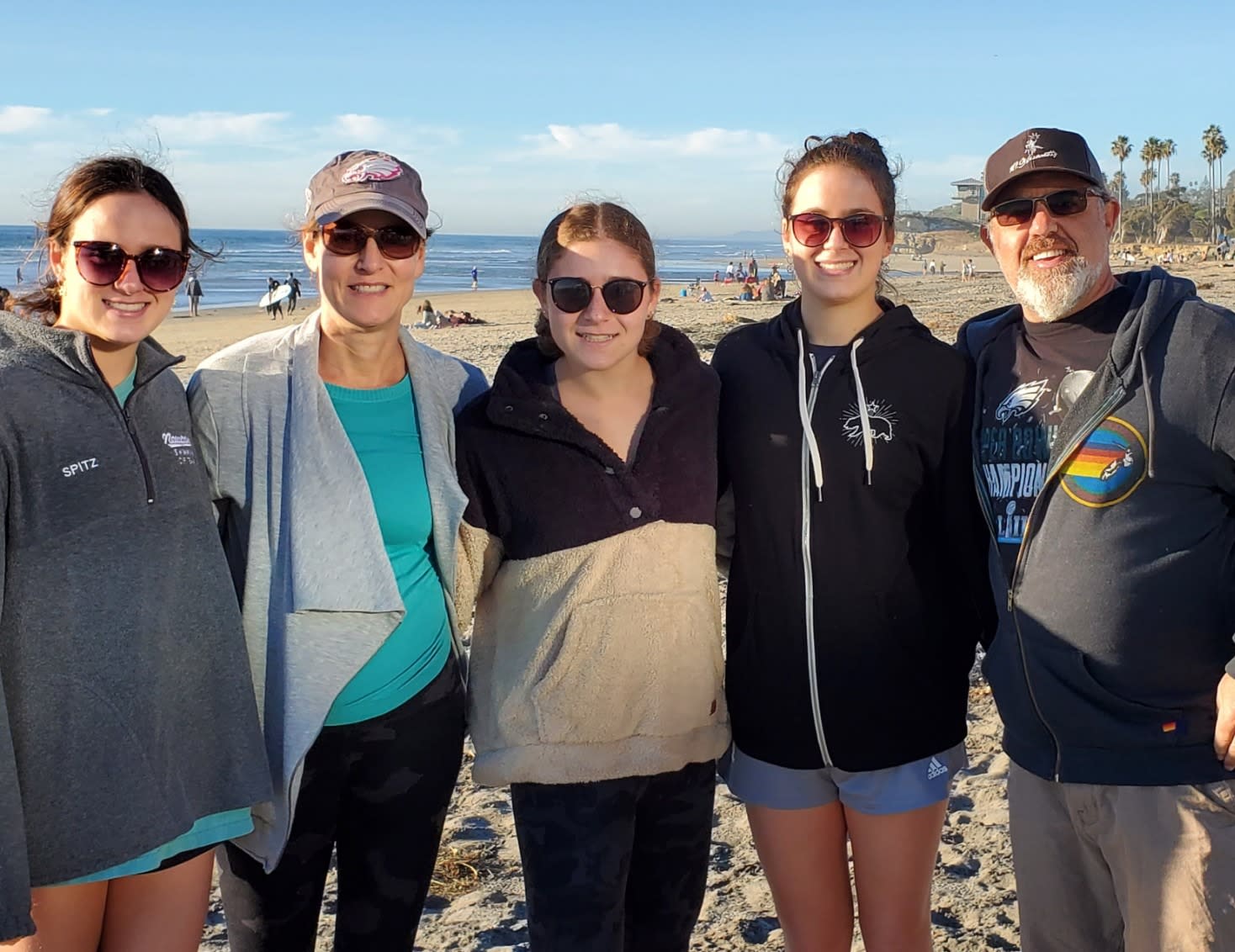 A group of five people including Jane, all wearing sunglasses, are standing on a beach with the ocean and palm trees in the background. Dressed casually in jackets and sweatshirts, they shine under the sun as they discuss their career journeys. Other beachgoers can be seen in the distance.
