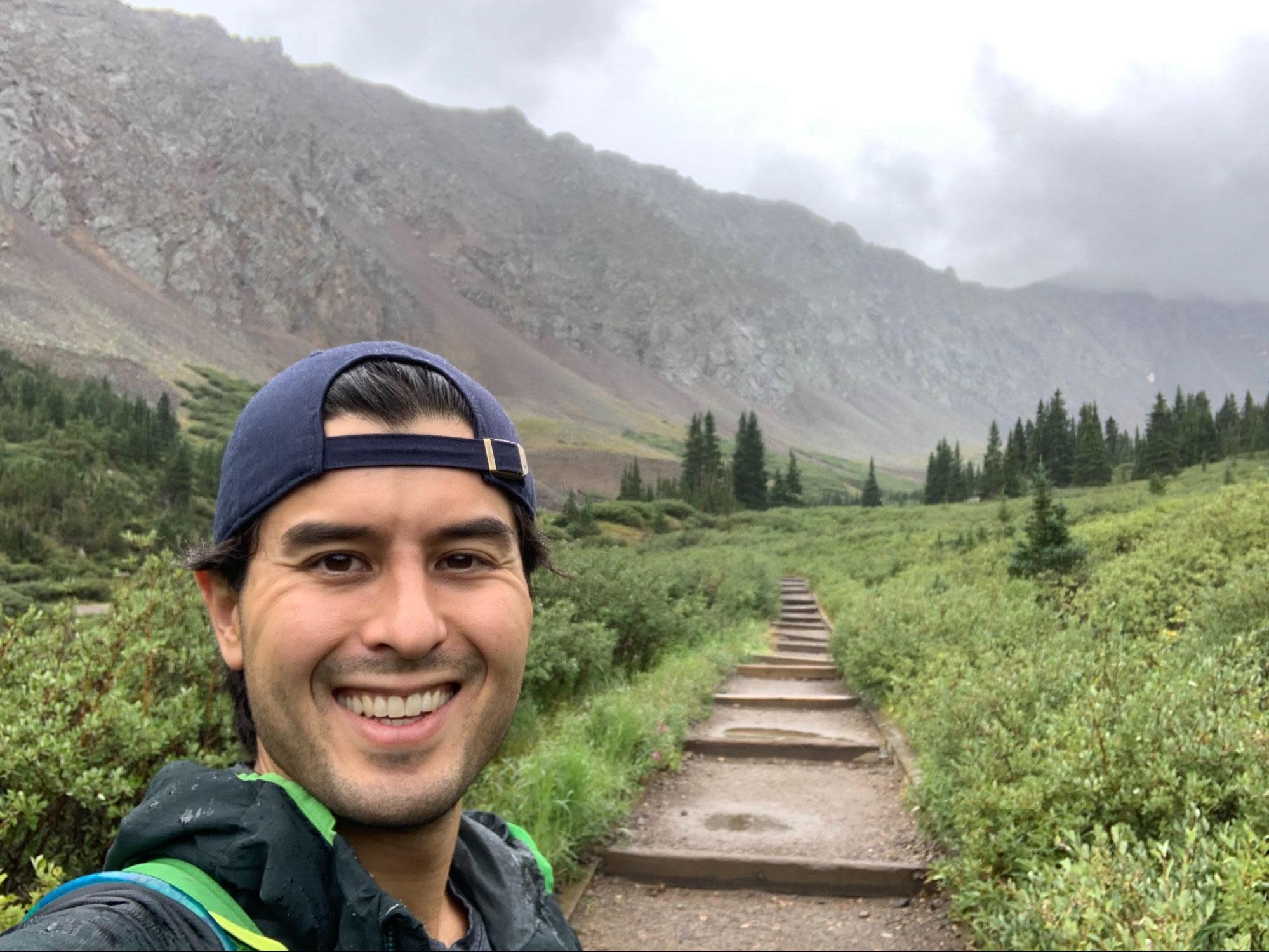 Keenan wearing a baseball cap and jacket is smiling for a selfie on a hiking trail, channeling his inner Ted Lasso. The background features a lush green landscape with a pathway leading into foggy, mountainous terrain. Evergreen trees dot the scene, and the sky is overcast.