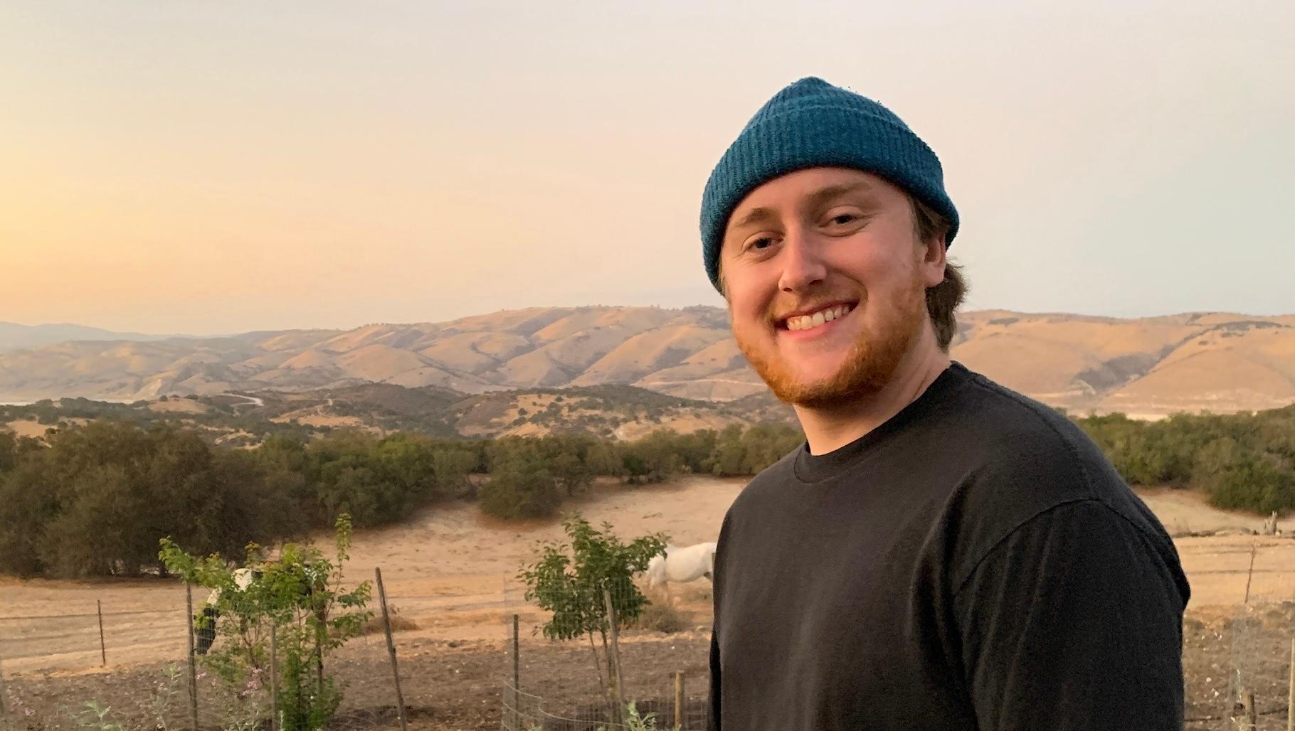 A person (Kevin) with a blue beanie and a black shirt smiles at the camera while standing outdoors. A scenic landscape featuring rolling hills, scattered trees, and a soft, cloudy sky unfolds behind them, reminiscent of the serene backgrounds in Avatar the Last Airbender.