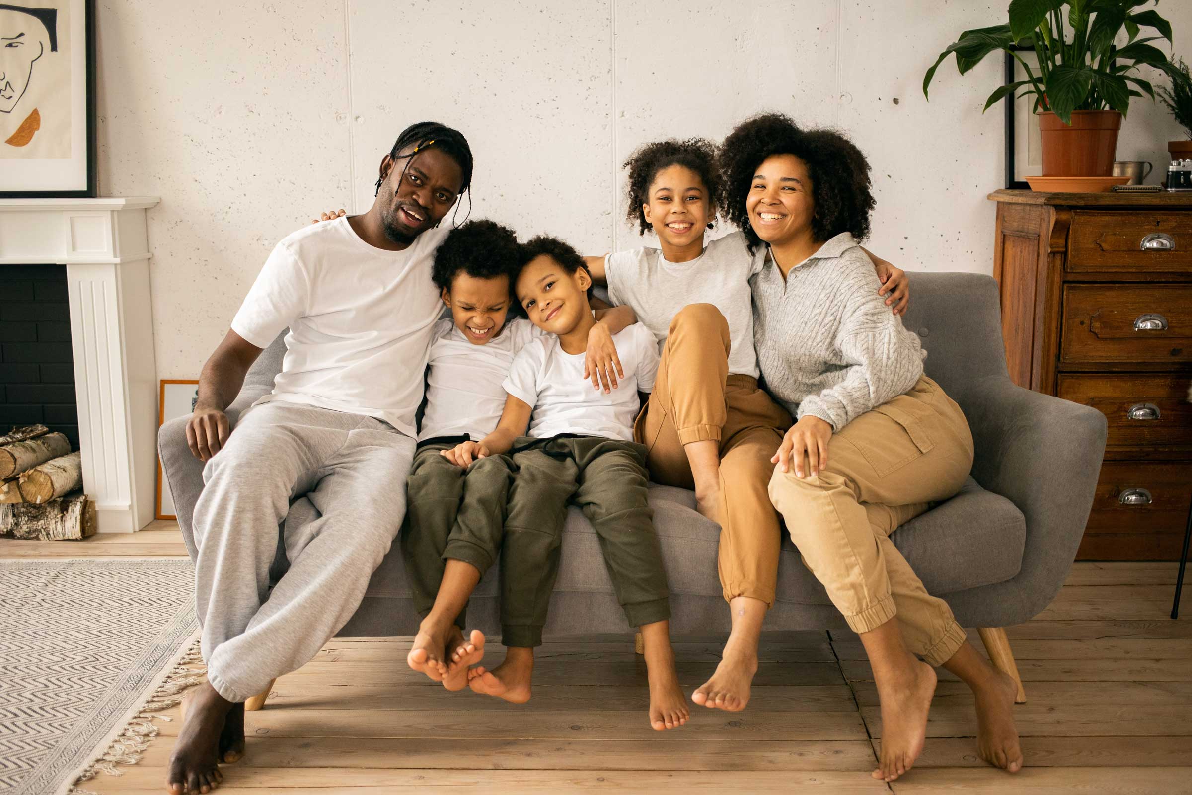 A family of five sits together on a grey couch, all smiling at the camera. The room has minimalistic decor with a plant on a wooden chest and artwork above a fireplace, embodying the love and support fostered through progressive policies like parental leave.