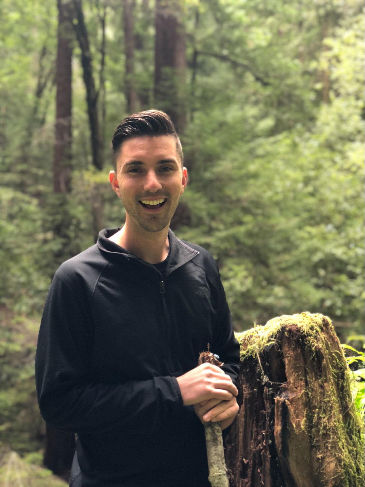 A person (Matt) with short dark hair, wearing a black zip-up jacket, stands smiling in a lush, green forest. They are holding a moss-covered tree stump with one hand, surrounded by tall trees and dense foliage.