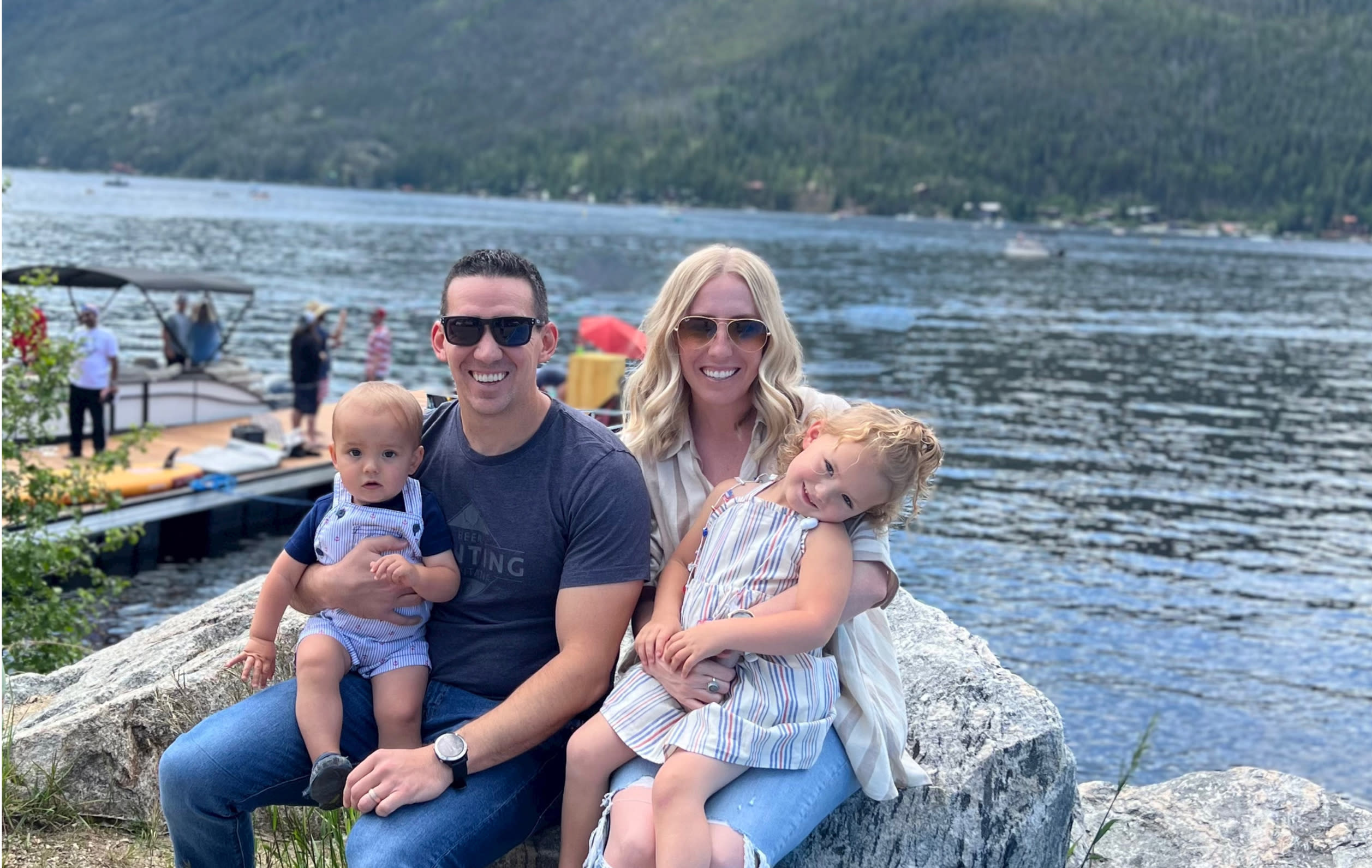 A family of four sits on a large rock by a lakeside. The father, wearing sunglasses and a dark t-shirt, holds a baby boy. The mother, with blonde hair and wearing sunglasses and a white top, holds a young girl who leans on her. Mountains rise in the background while they discuss remote work culture.