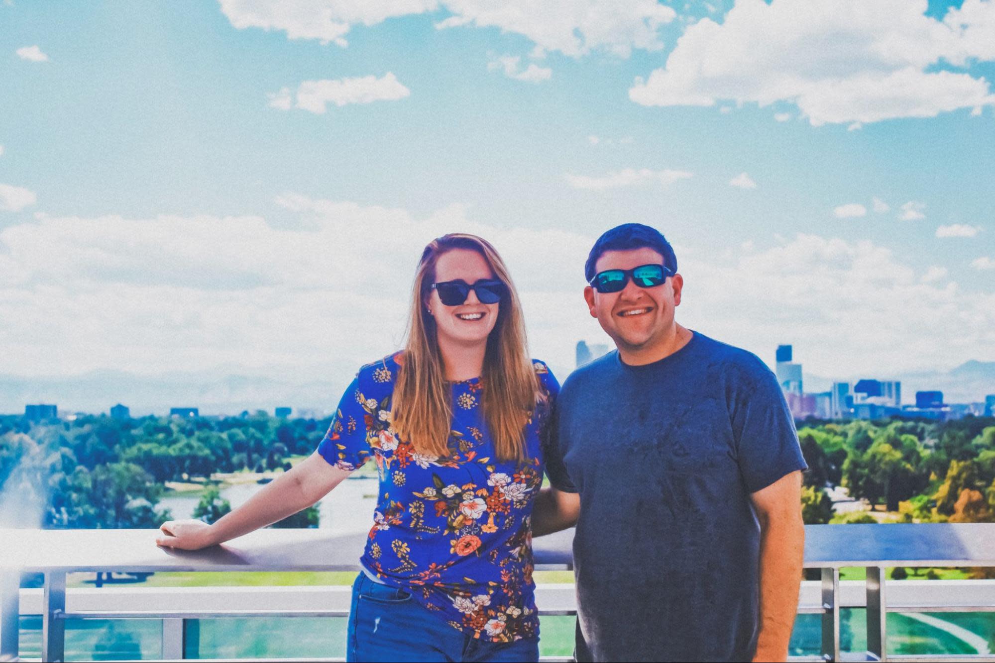 Two people stand on a sunny outdoor balcony with a vibrant cityscape and green park in the background. They are smiling and wearing sunglasses. The person on the left sports a blue floral shirt, while Mike is wearing a dark gray shirt.