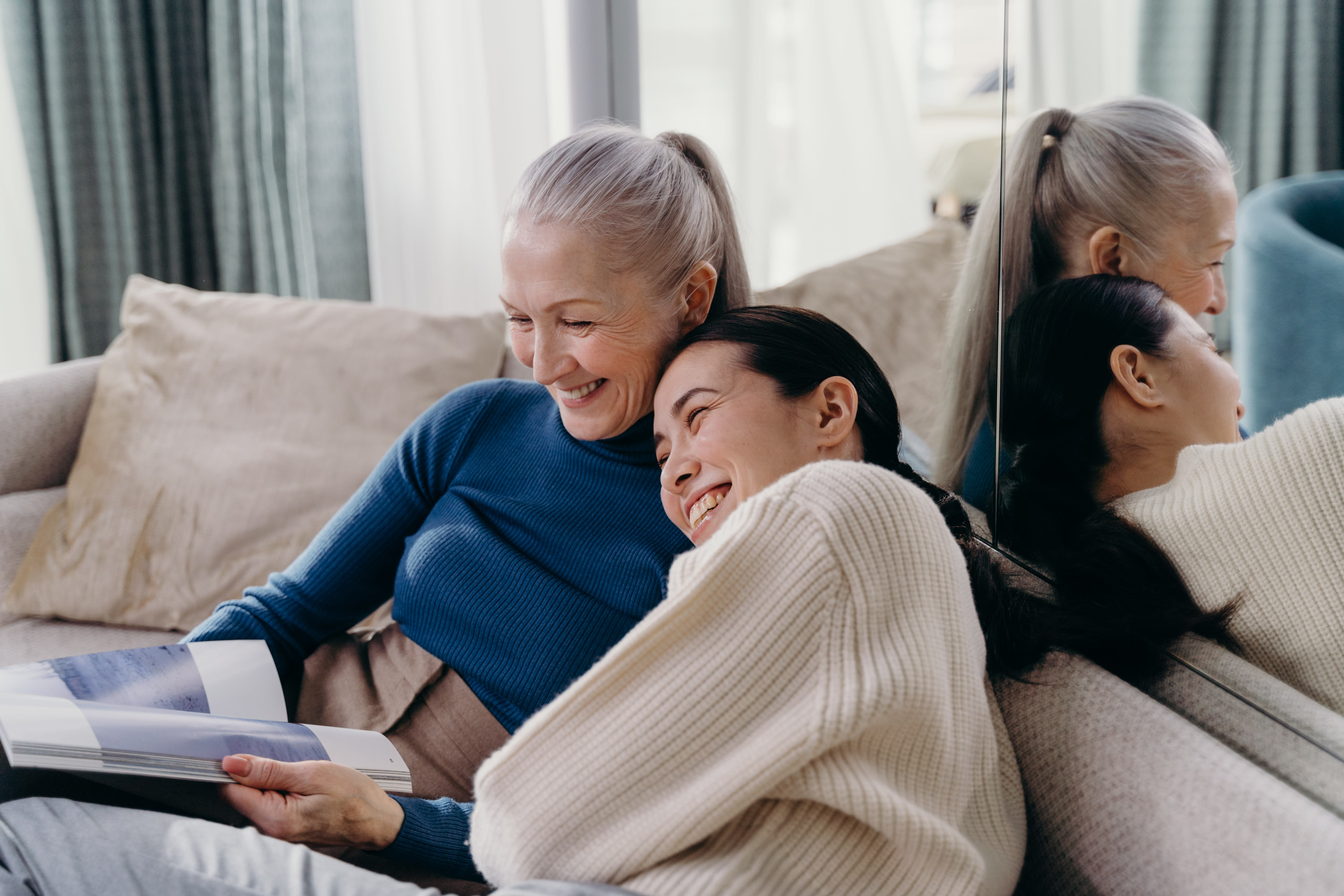 Two women sit closely together on a couch, smiling and looking at a book. The woman on the left has gray hair in a ponytail and wears a blue long-sleeve shirt. The woman on the right, demonstrating personal bonds in family leave, has dark hair in a ponytail and wears a cream sweater, leaning her head on the other's shoulder.