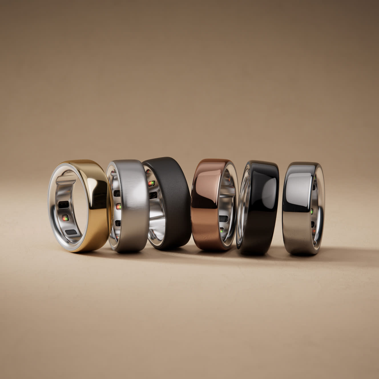 A row of six smart Oura Rings in different metallic finishes, including silver, gold, black, brown, and matte grey, displayed on a neutral background. Each ring has a sleek, modern design.