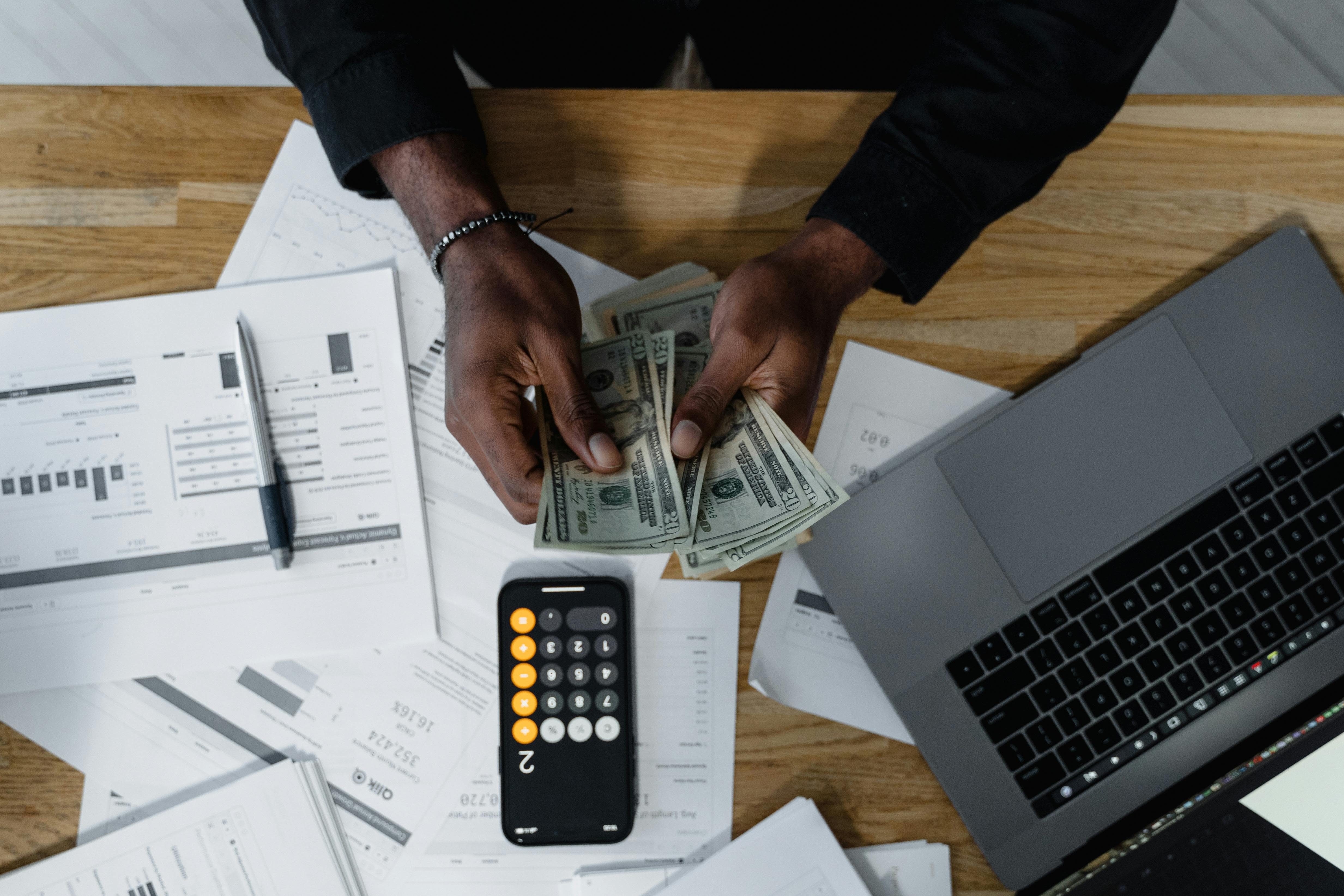 A person is counting U.S. dollar bills at a wooden desk, surrounded by financial documents detailing employer policy. The desk also holds a pen, calculator, and open laptop. Numbers flash on the calculator while papers are scattered around.
