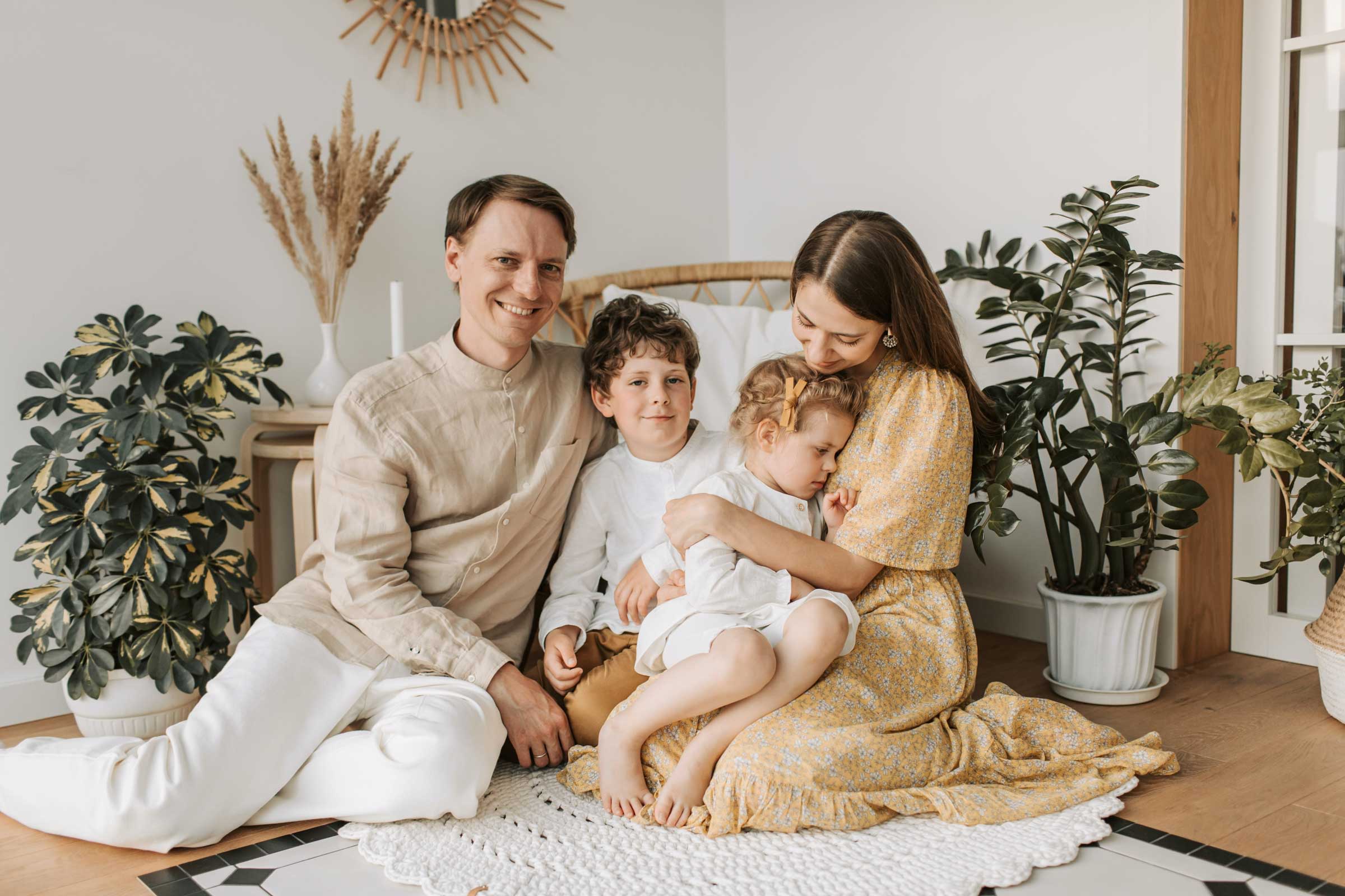 A family of four sitting on the floor in a bright, cozy room with plants. The father sits on the left, smiling. The mother, in a floral dress, holds their two children—one sitting and one resting on her lap. The atmosphere is warm and affectionate, a perfect example of Minnesota paid family leave in action.