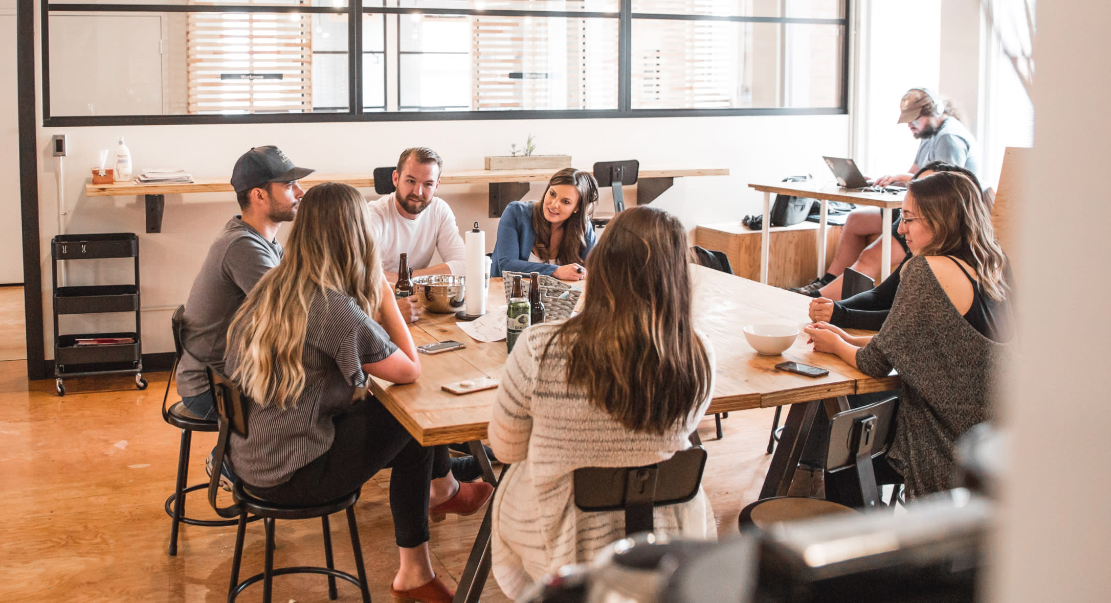 A group of seven people sit around a wooden table in a bright, modern office. They are engaged in discussion, with some leaning forward and others sitting back. In the background, a person works on a laptop at a separate desk. The atmosphere seems collaborative and infused with improv and humor.