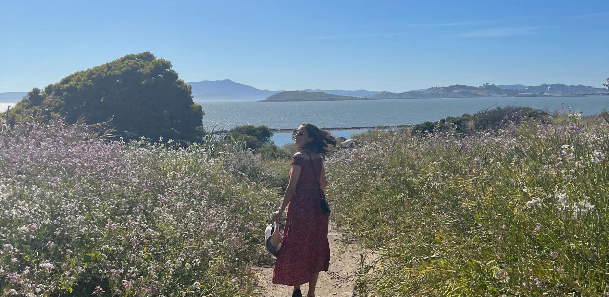 A person wearing a red dress and holding a hat stands on a dirt path surrounded by tall grass and wildflowers. They are facing a scenic view of hills and a body of water under a clear blue sky, embodying the essence of women in tech advocacy with their confident and curious stance.