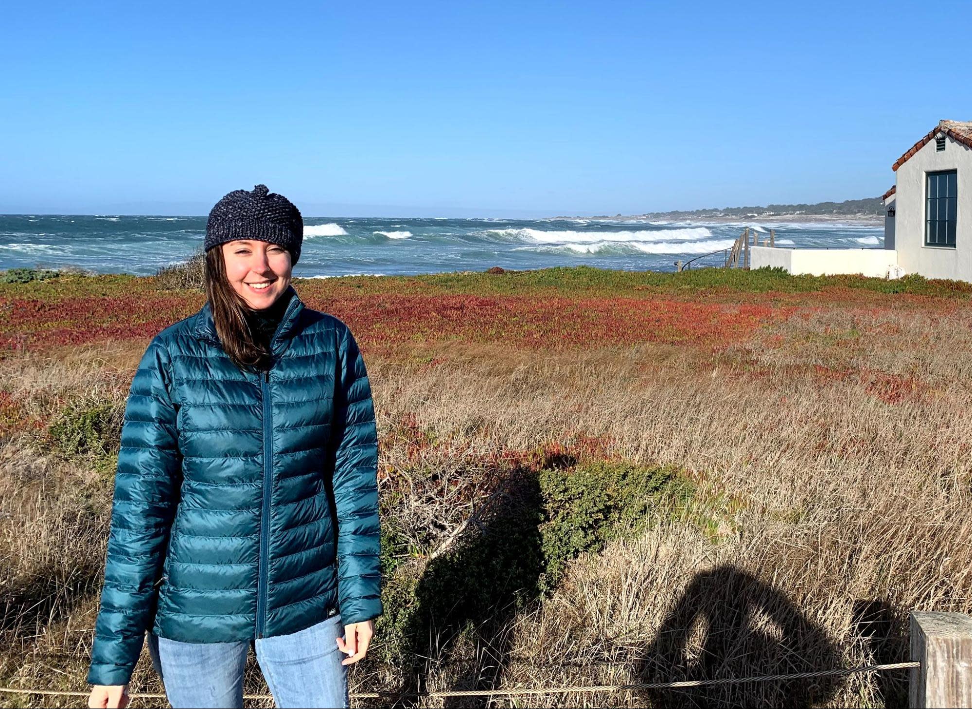 Sierra wearing a blue puffer jacket and knit hat stands smiling in front of a coastal landscape with red and brown vegetation. The ocean waves crash behind her under a clear blue sky. A building is visible to the right while a fence casts shadows on the ground.