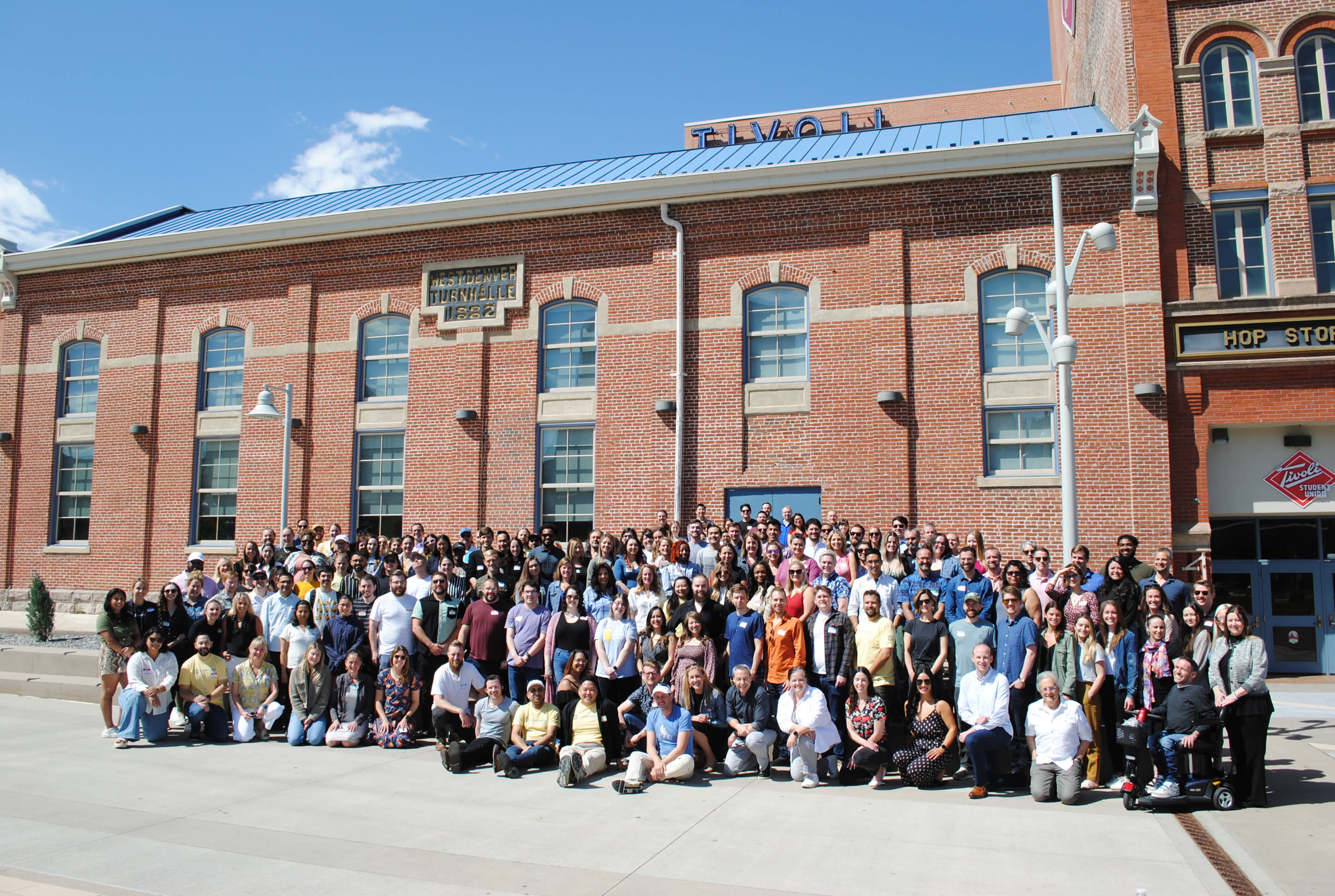 A large group of people poses together in several rows for an outdoor photo in front of a historic red brick building with arched windows and a blue sign that reads "Tivoli." The sky is clear and blue, with a few scattered clouds.