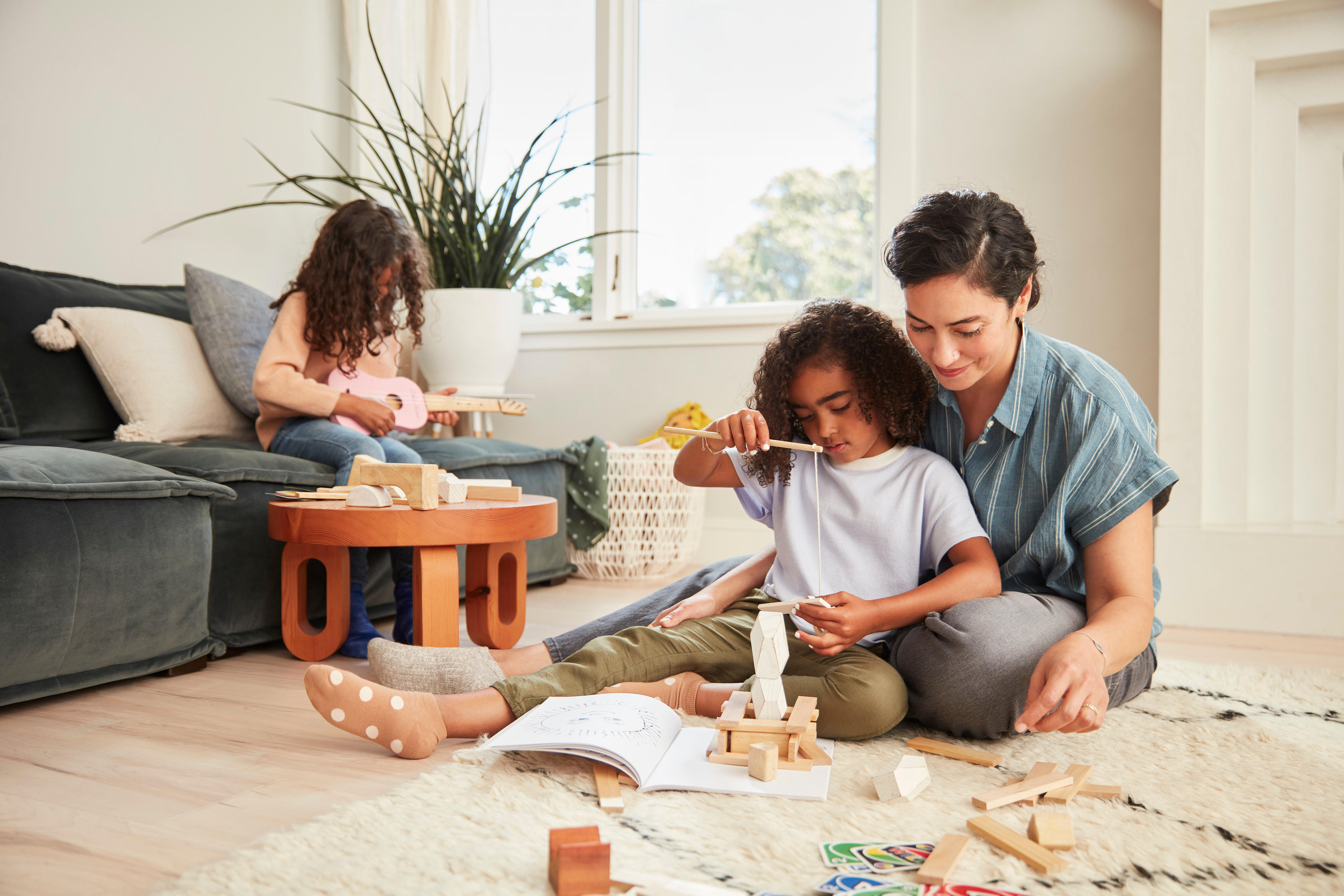 A woman sits on the floor helping a young child build with wooden blocks. Another child is sitting on a couch in the background, engaged in an activity. The scene is set in a cozy, well-lit living room with a plant near the window, reflecting modern family structures.