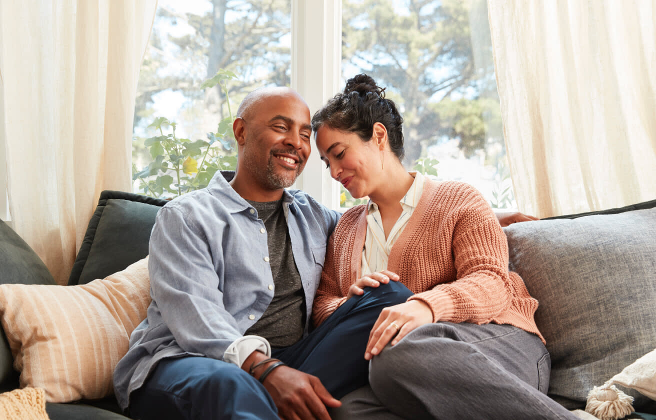 A smiling couple sits closely together on a cozy sofa in a sunlit room with large windows. The man, in a blue shirt and gray t-shirt, has his arm around the woman, who is wearing a peach cardigan and white blouse, resting her head on his shoulder, creating an idyllic moment free of legal risks.