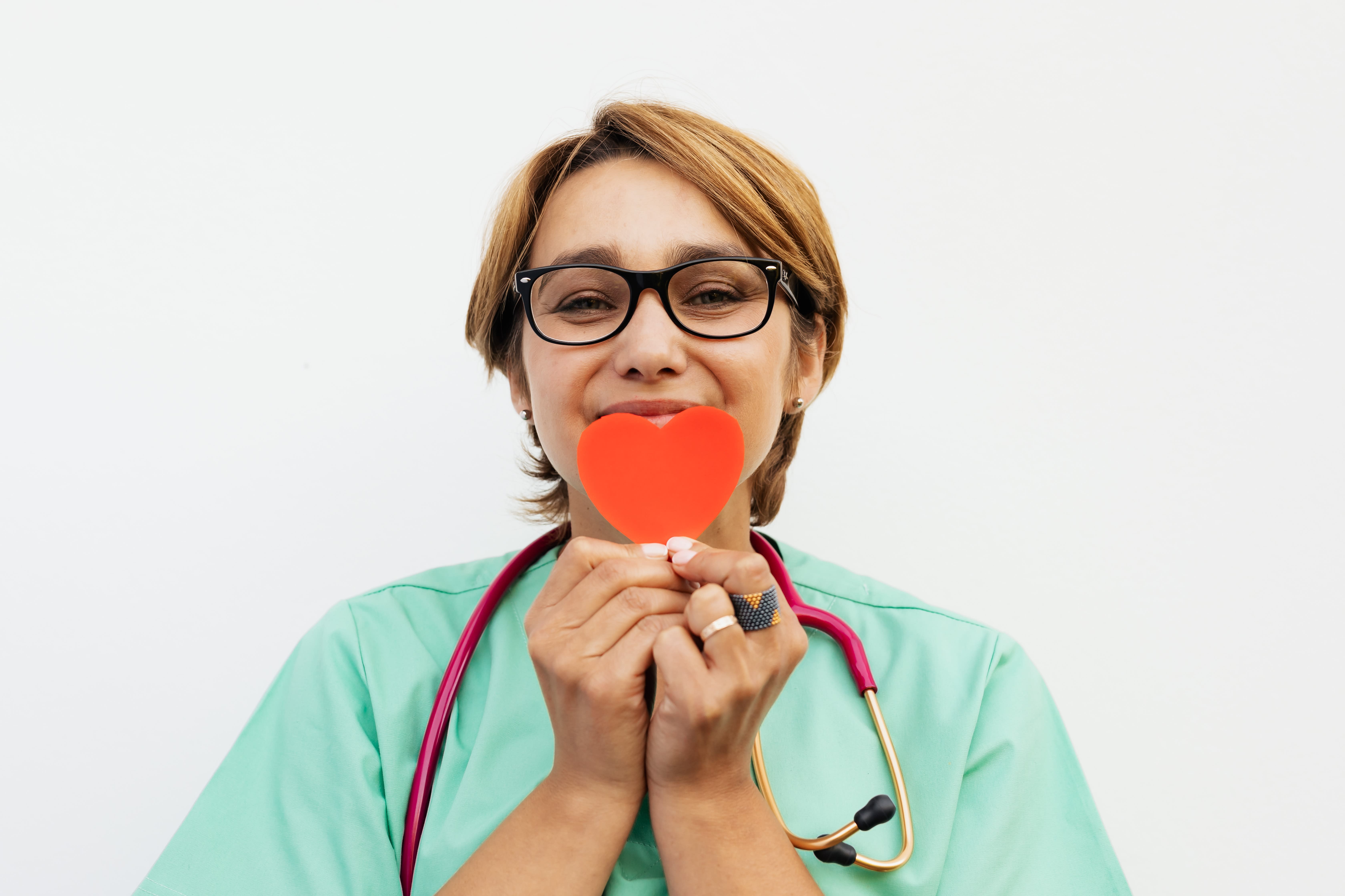 A healthcare professional wearing green scrubs, a stethoscope around the neck, and glasses holds up a red heart shape in front of their face, smiling gently. The background is plain white, highlighting their commitment to promoting organ donation leave.