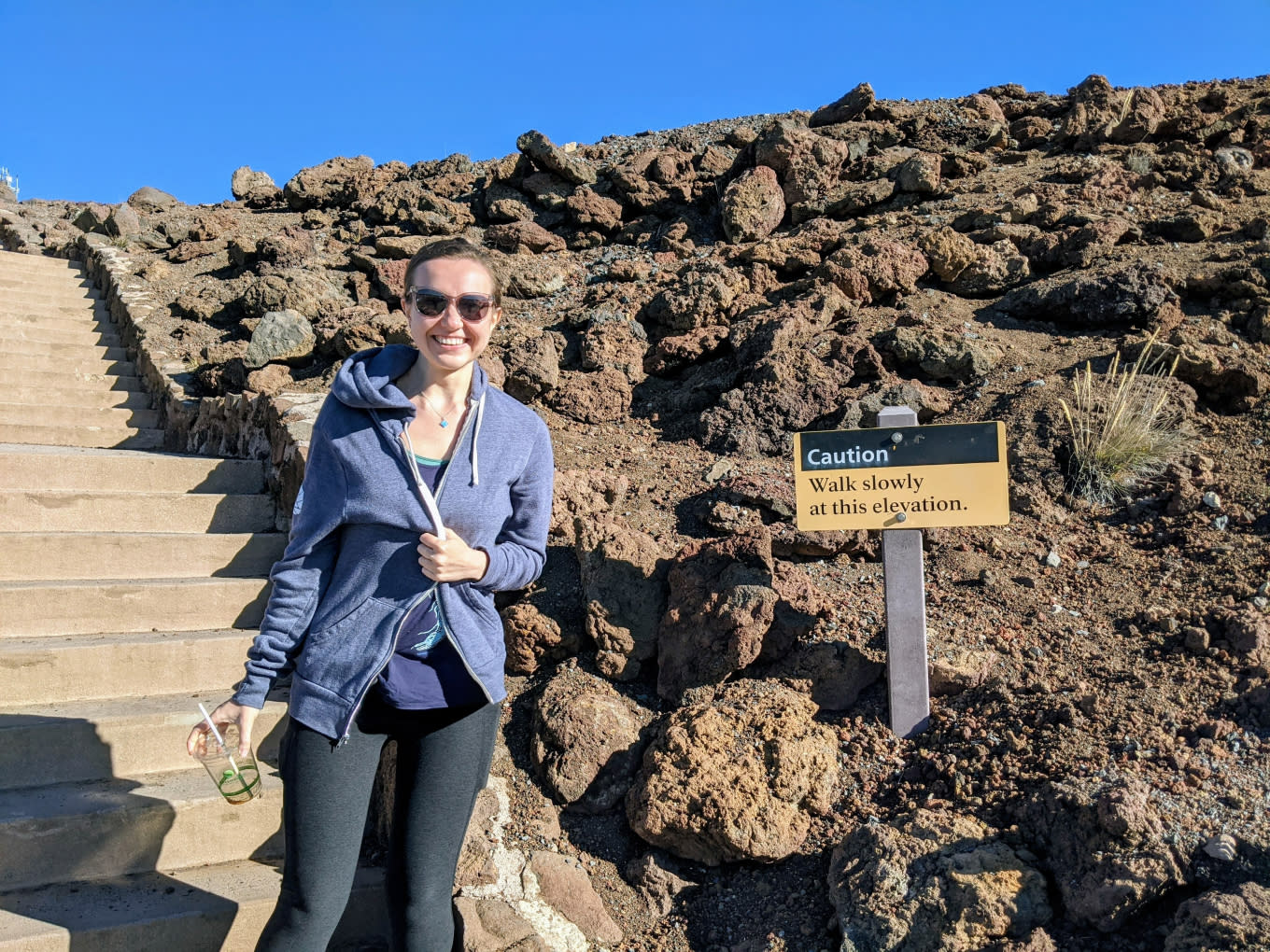 A person wearing a blue hoodie and sunglasses stands next to a rocky path. A sign beside them reads "Caution: Walk slowly at this elevation." They are holding sunglasses and smiling, with a staircase visible to the left. The sky is clear and blue, mirroring their enthusiasm for meaningful work.