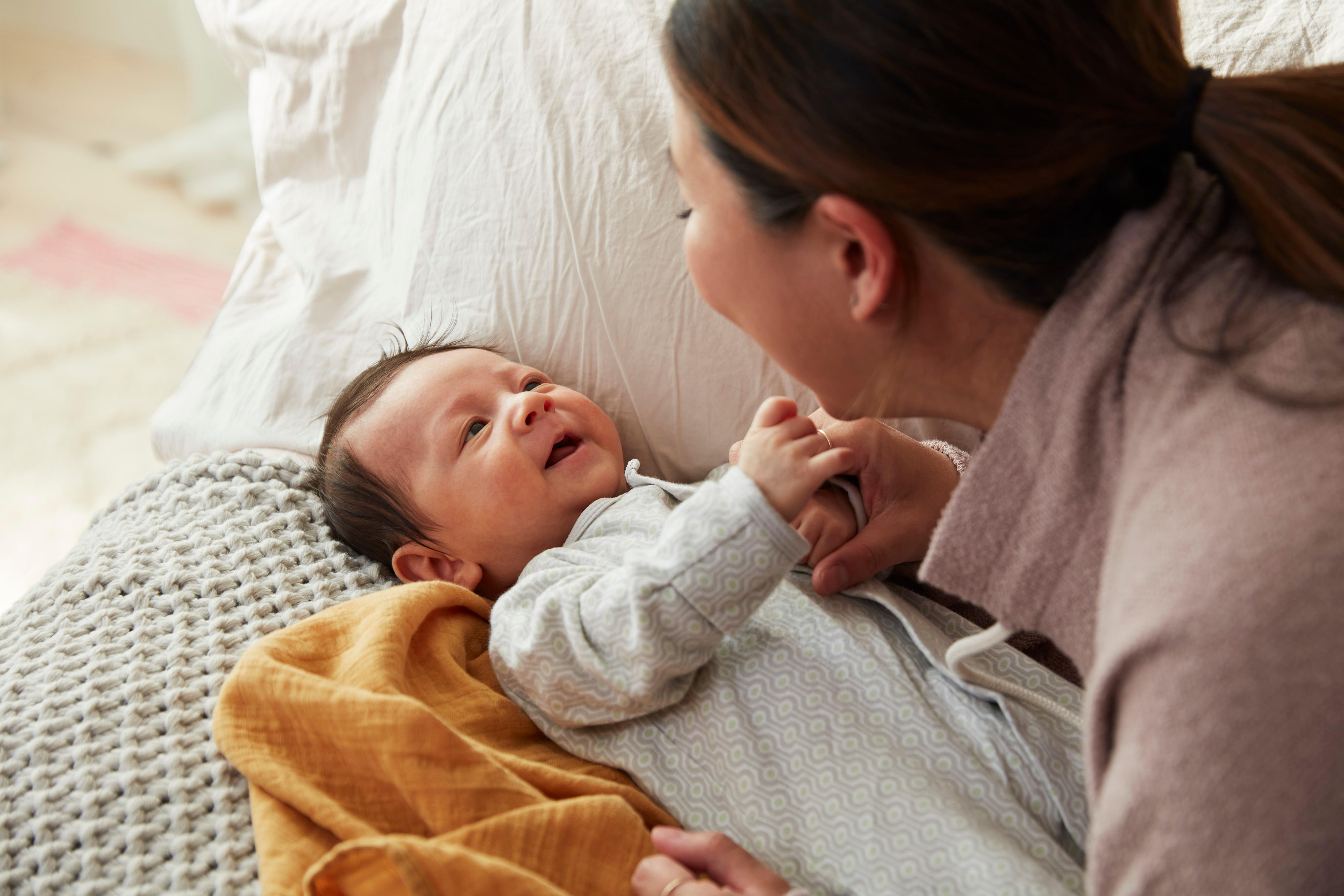 A caregiver smiles and interacts with a baby lying on a bed. The baby looks up and holds the caregiver's hand while wearing a white outfit. The bed is covered with a textured blanket, and a yellow cloth is draped nearby, illustrating the warmth often celebrated in New York's expanded family definition.
