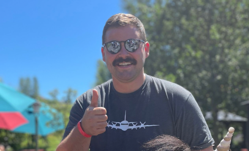 A man with a mustache and sunglasses gives a thumbs-up. He is wearing a gray t-shirt with an airplane graphic, standing outdoors with a bright, sunny sky and green trees in the background. Colorful umbrellas are visible behind him, hinting at his recent La Jolla Cove snorkeling adventure.