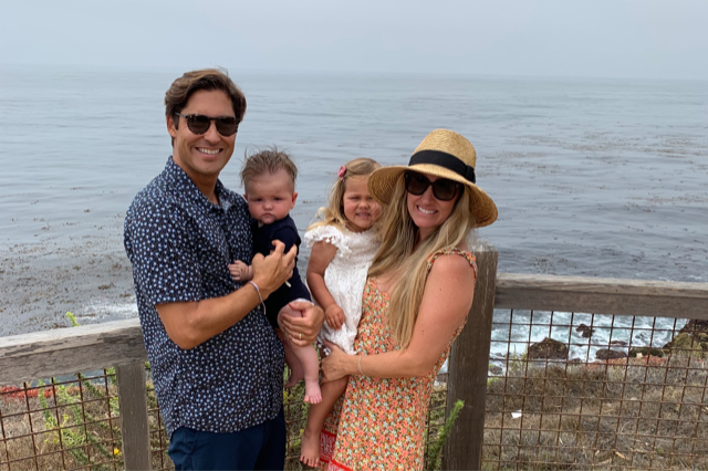 A man and woman (Christa) stand by the ocean, smiling and holding two young children. The man, wearing sunglasses and a patterned shirt, holds a baby. The woman, in a floral dress and sunhat, holds a toddler in a white dress. 